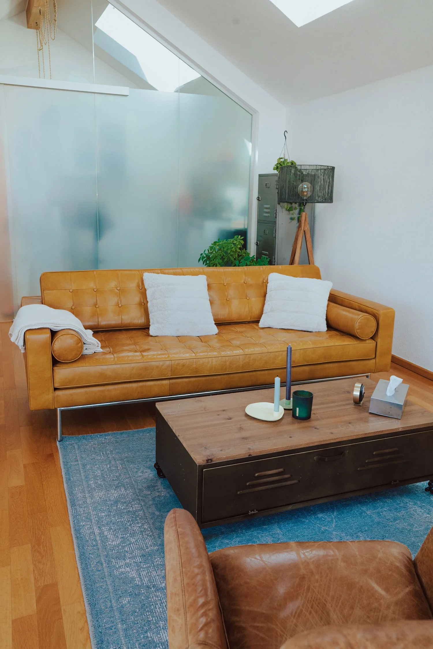 Gemütlicher Praxisraum mit braunen Ledersofas, weißen Kissen, einem Holztisch mit Kerzen und Stofftaschentüchern, grünem Pflanzen und moderner Wandkunst. Es handelt sich um den Hauptraum der Praxis in Trier.