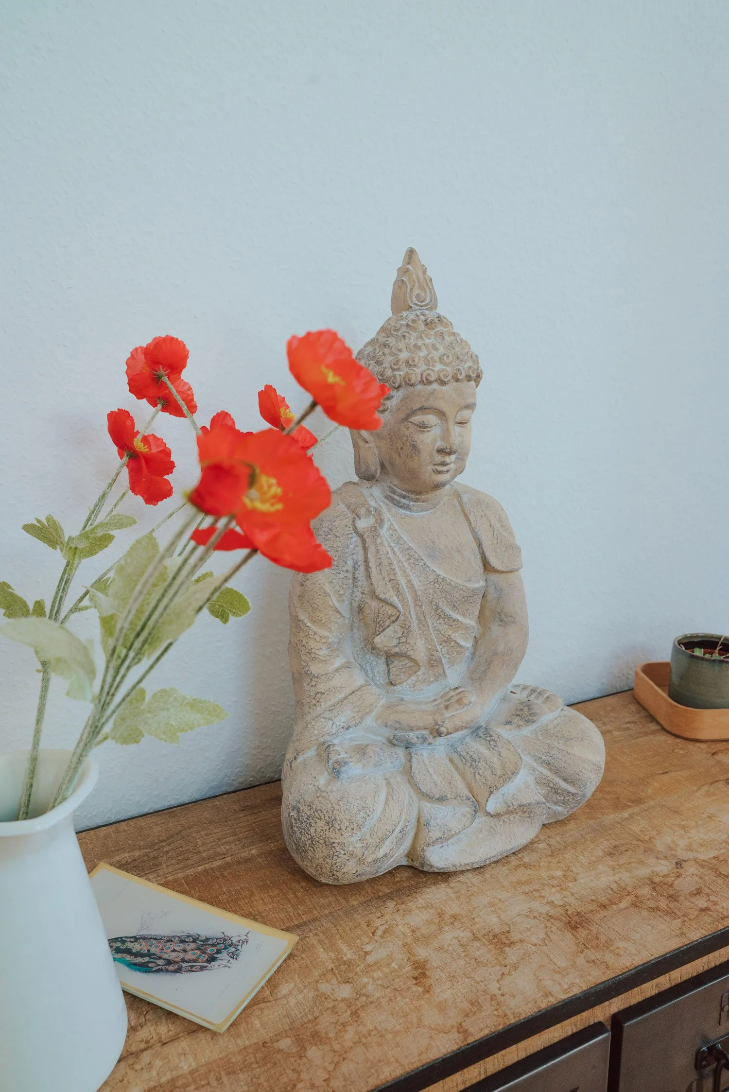 Eine Keramikfigur eines sitzenden Buddha auf einem Holzsideboard, daneben eine Vase mit roten Mohnblumen und eine kleine Schale vor einer weißen Wand.