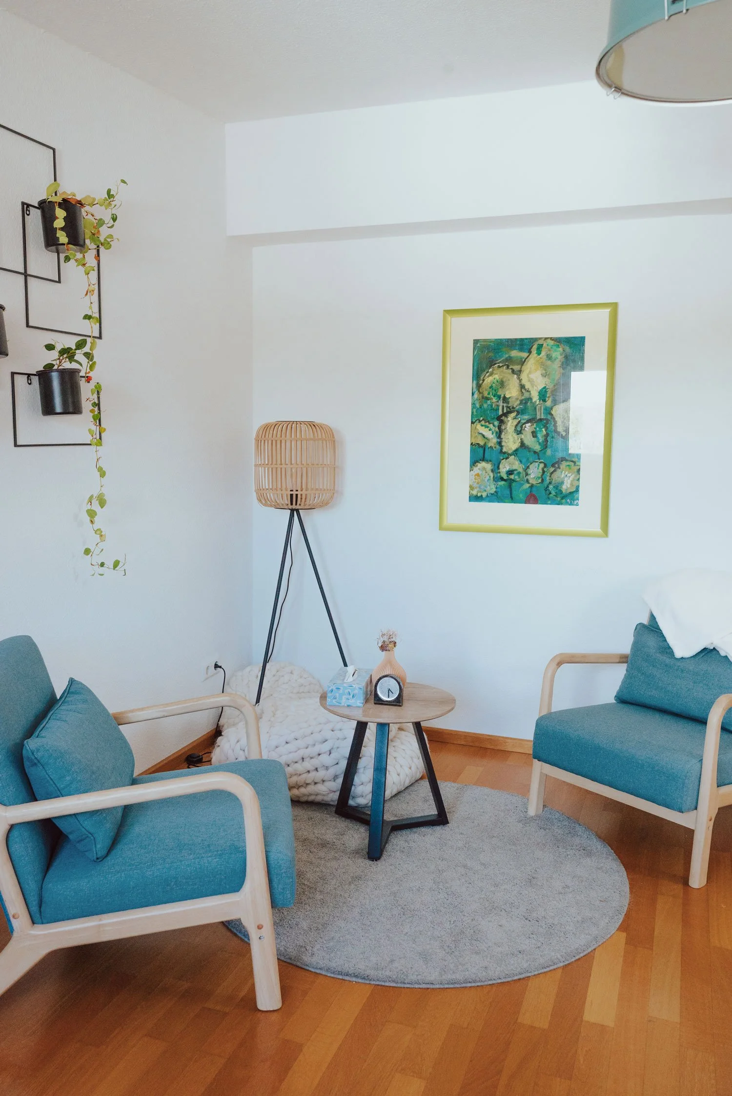 Gemütliche kleiner Praxisraum mit zwei blauen Sesseln und einem runden Holztisch mit Taschentuchbox und Deko. Im Hintergrund steht ein Stehlampe mit einem Rattan-Schirm, an der Wand hängen Kunstwerke und Pflanzen. Ein Teppich liegt auf Holzboden.