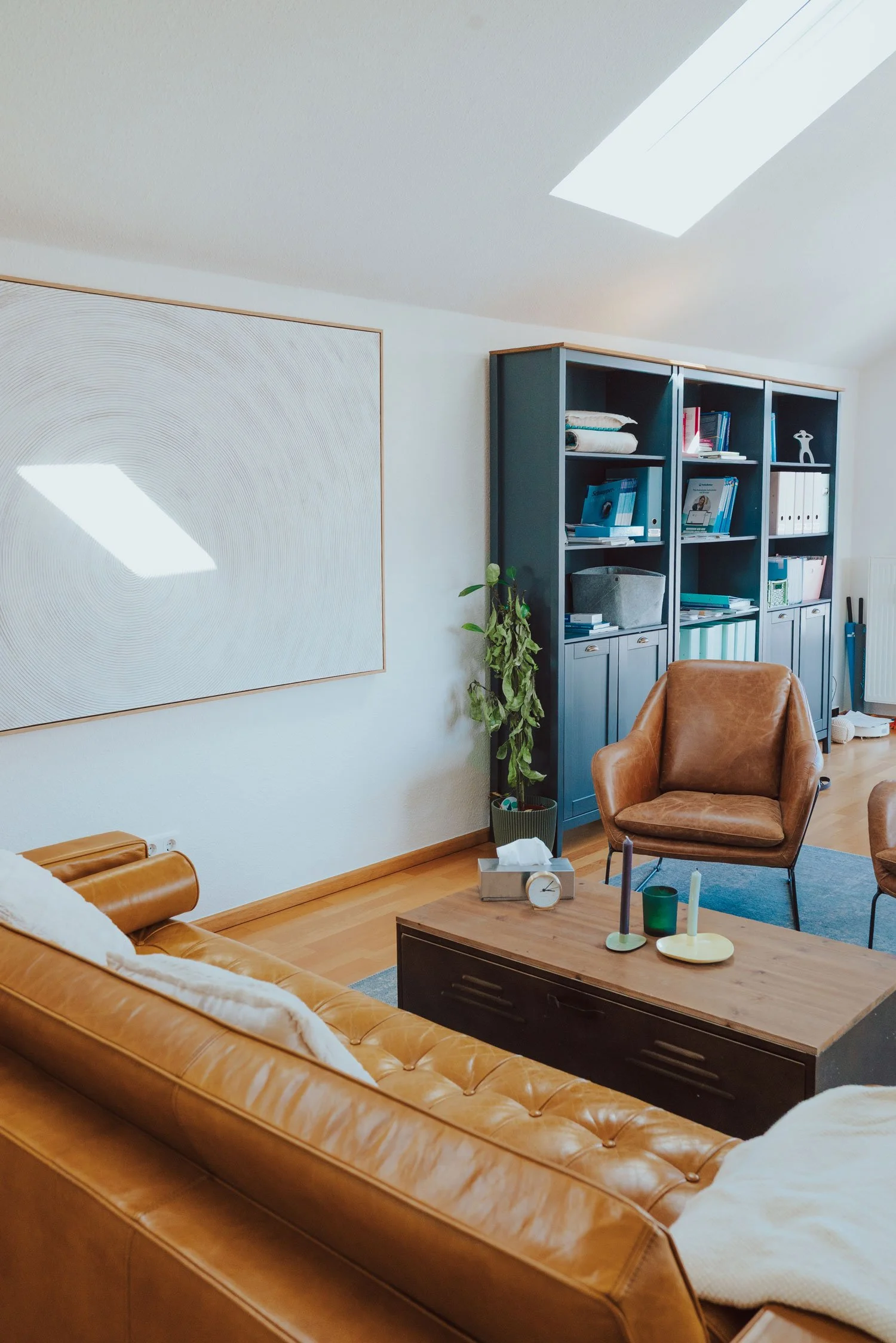 Hauptrraum der Praxis Trier mit braunem Ledersofa, einem Sofatisch mit Kerzen und Dekoration, einem blauen Bücherregal, einem braunen Sessel und Pflanzen.