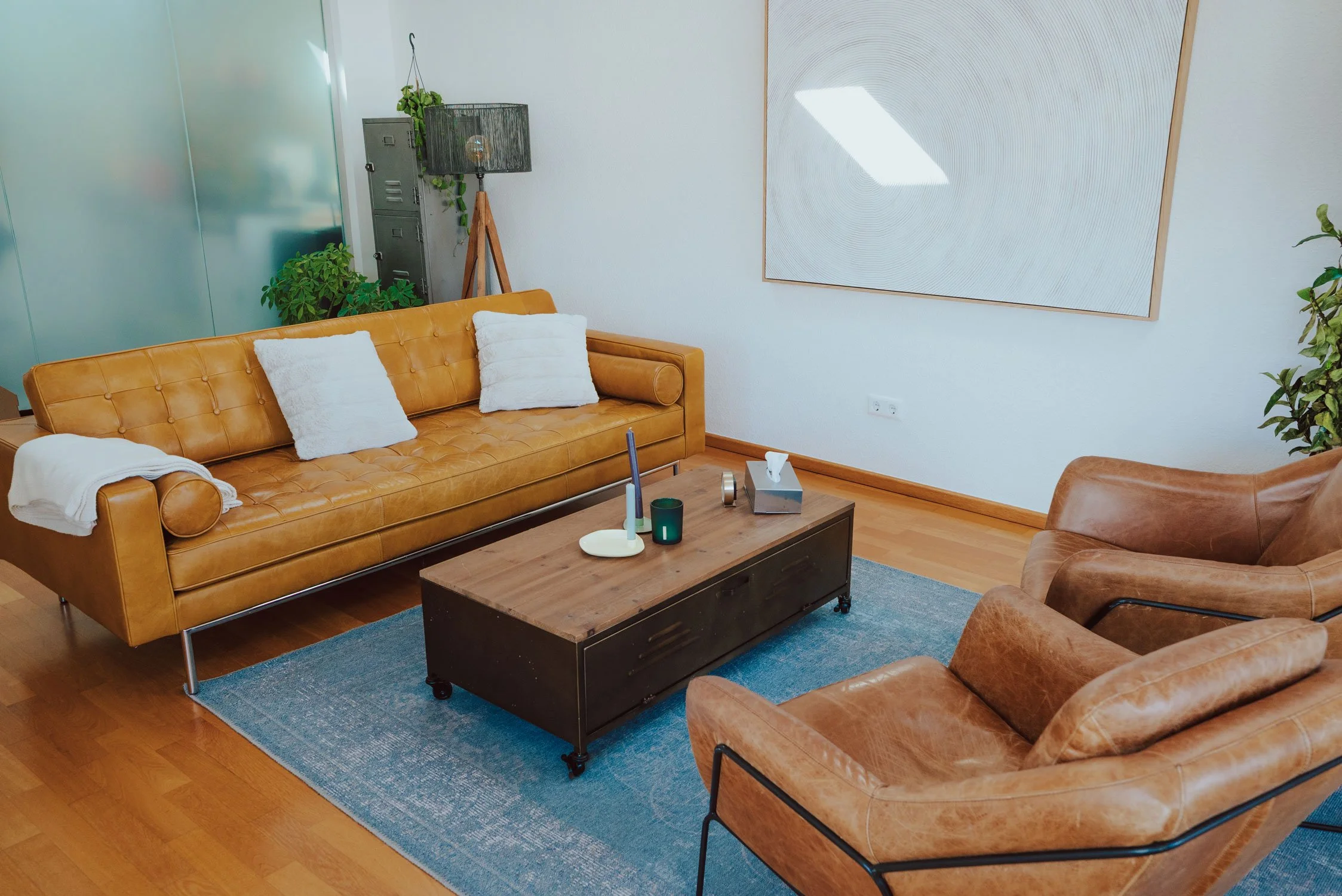Gemütlicher Praxisraum mit braunen Ledersofas, weißen Kissen, einem Holztisch mit Kerzen und Stofftaschentüchern, grünem Pflanzen und moderner Wandkunst. Es handelt sich um den Hauptraum der Praxis in Trier.