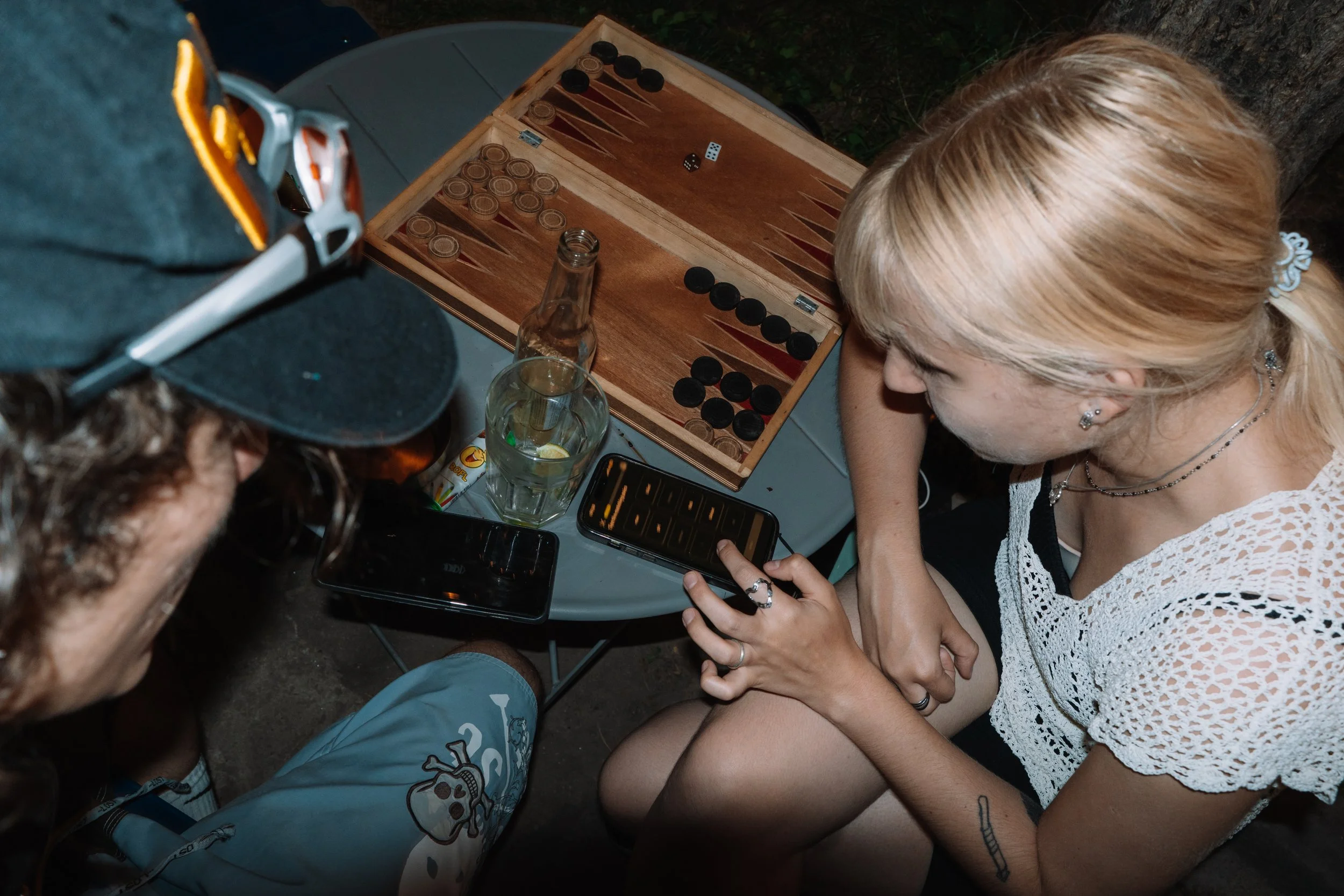 A woman with blonde hair and earrings playing backgammon at a table outdoors at night, with a young man partially visible in the foreground. There are drinks and a phone on the table.