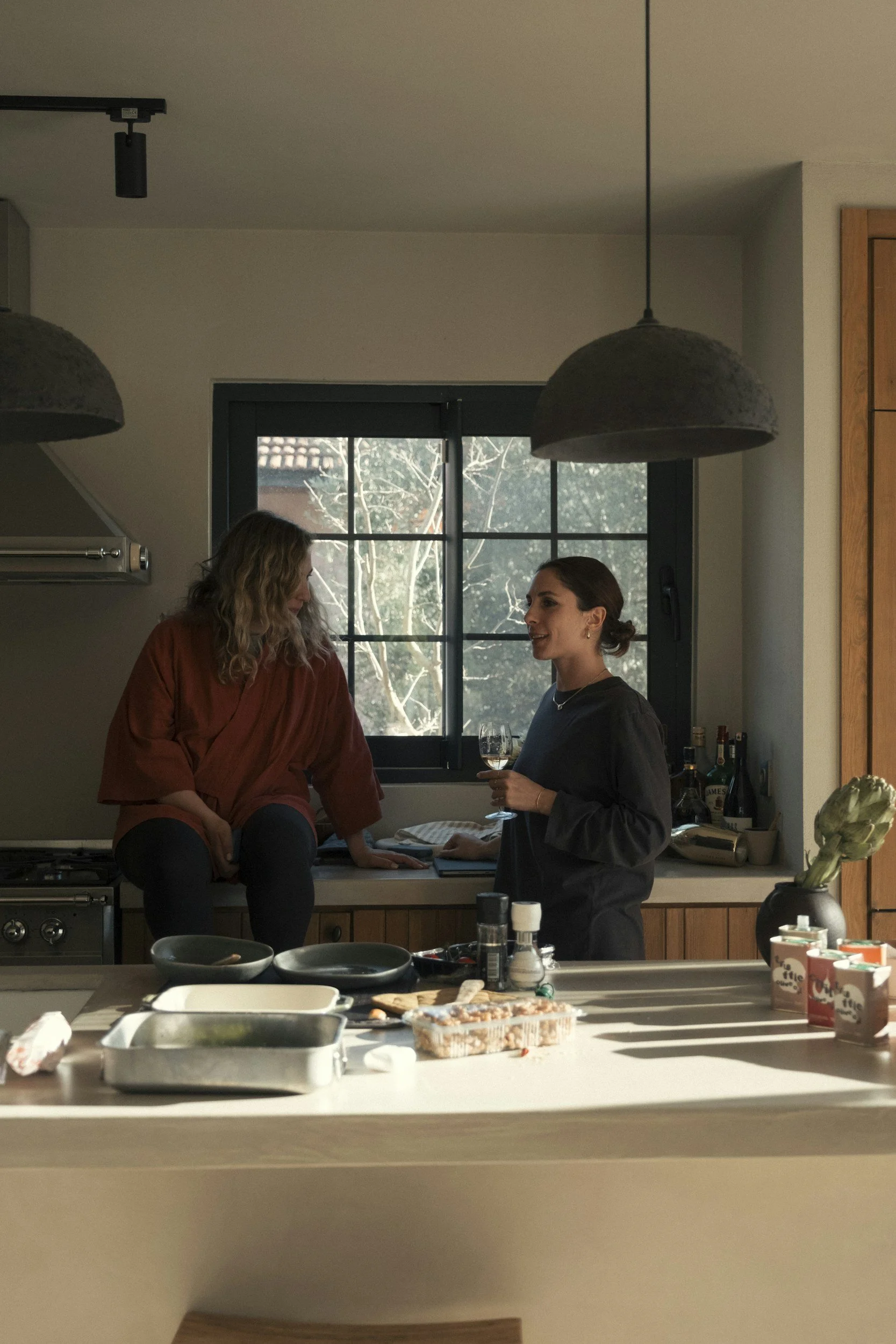 Two women having a conversation in a kitchen with hanging lights, window, and various kitchen items on the counter.