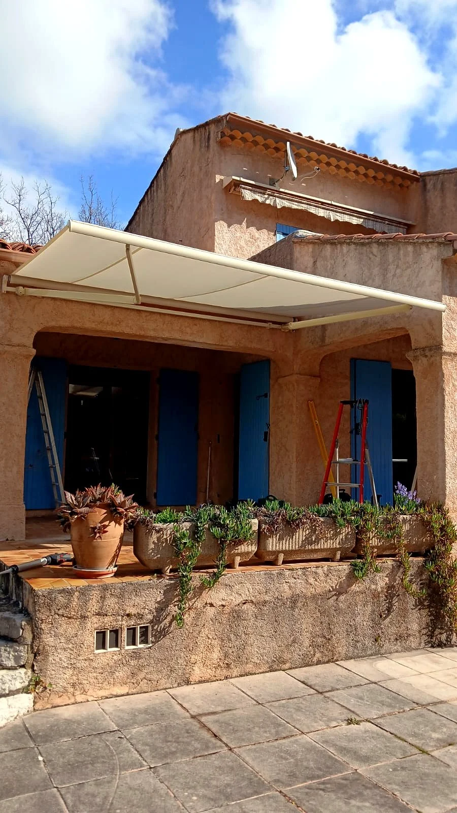 Store banne écru déployé devant une façade en pierre à volets bleus