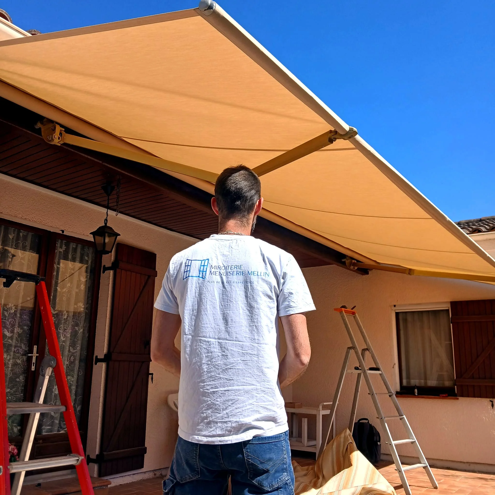 Technicien de la Miroiterie Menuiserie Mellin posant un store banne beige au-dessus d’une terrasse