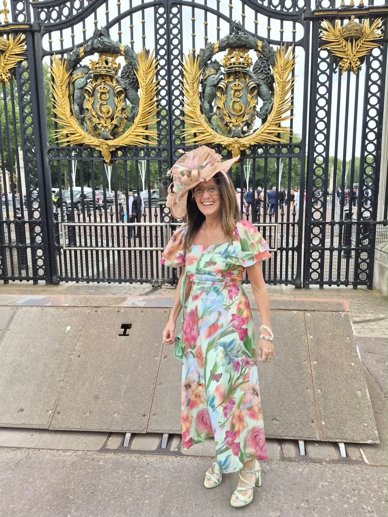 A woman wearing a floral dress and large pink hat smiling in front of a black gate with gold accents and emblem, with people and trees in the background.
