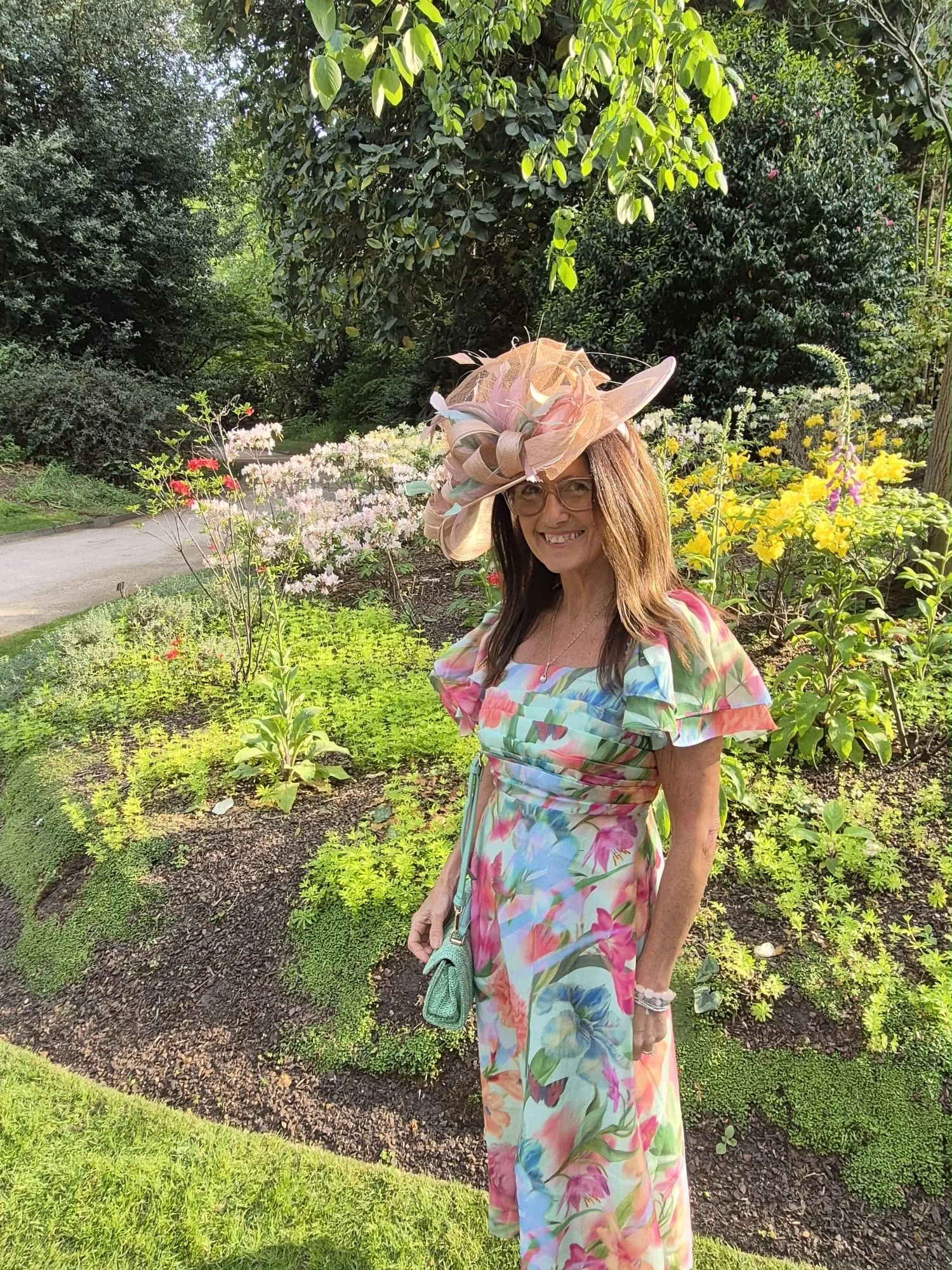 A woman standing in a garden with various colorful flowers, wearing a floral dress and a large, elaborate pink hat with decorative elements, smiling at the camera.