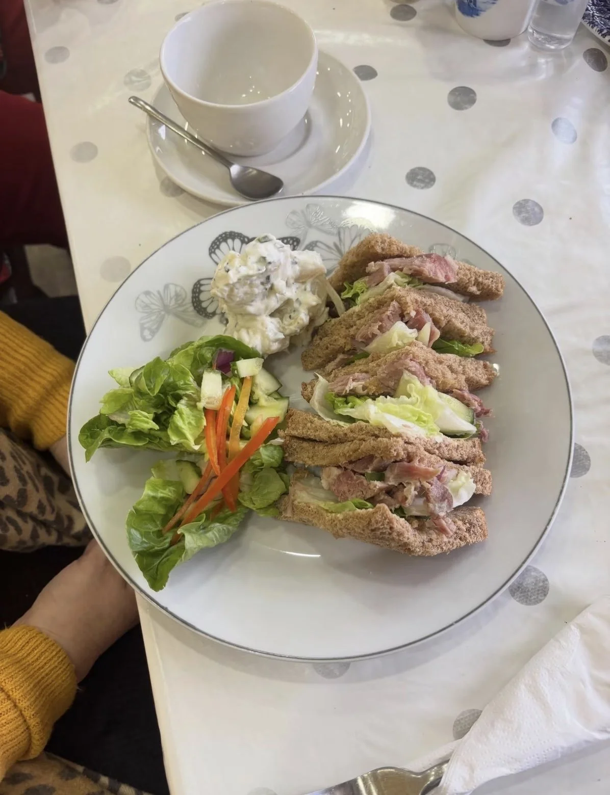 A plate with three ham and lettuce sandwiches, a side salad with lettuce, carrots, and cucumbers, a scoop of potato salad with chopped onions, and an empty white bowl with a saucer and spoon.