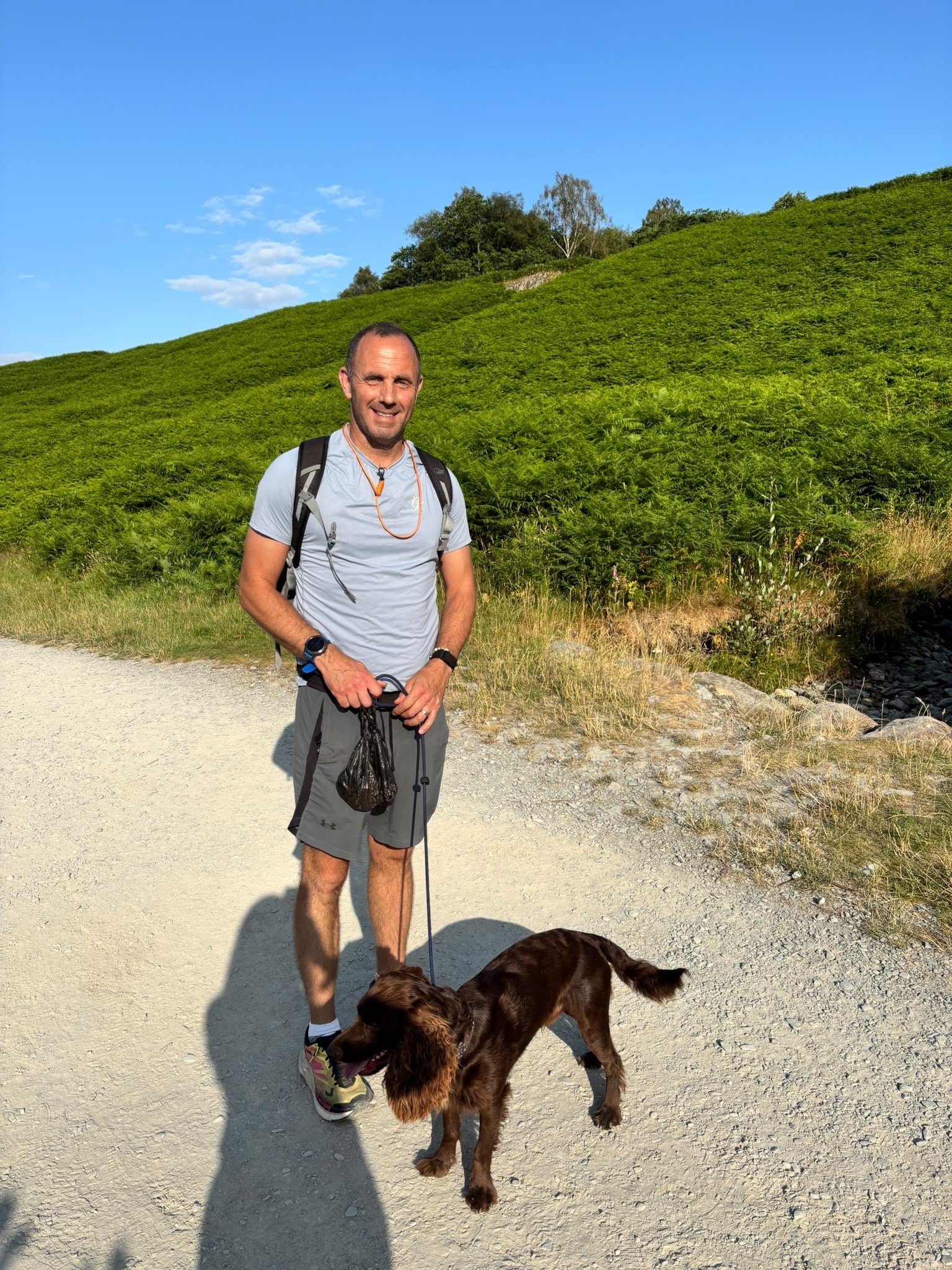 A man smiling while standing on a dirt trail with a brown dog on a leash in front of a green hillside and blue sky.