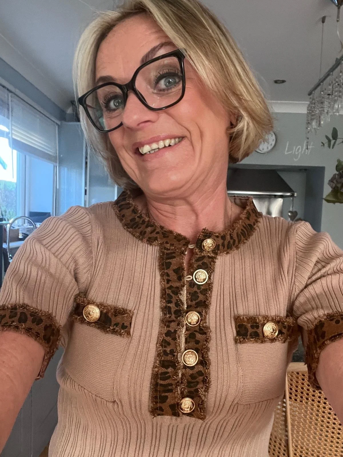 A woman with blonde, shoulder-length hair and glasses takes a selfie inside a house. She is smiling and wearing a beige top with leopard print accents and gold buttons.