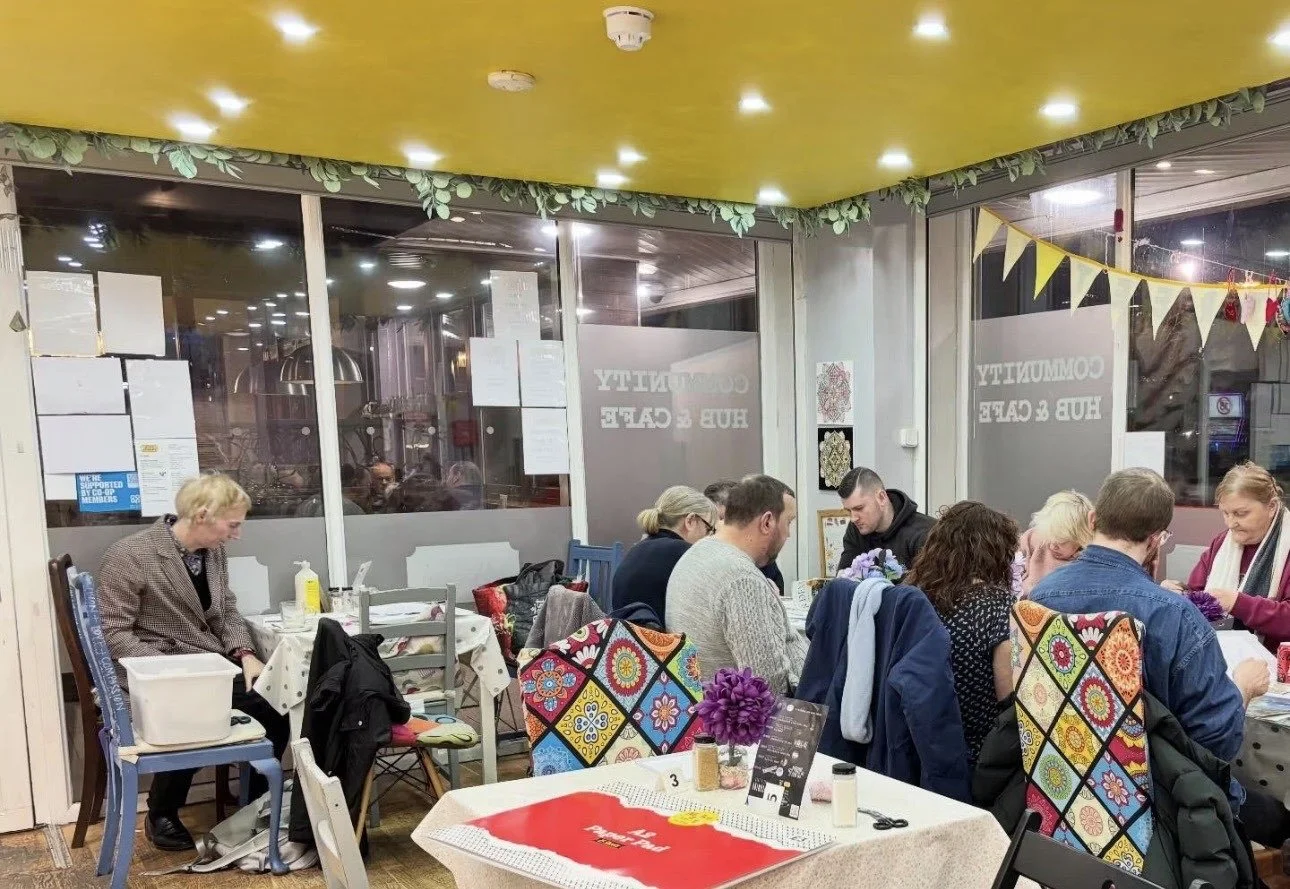 Group of people sitting at a table in a colorful cafe, with floral chair covers and window decals reading 'Community Hub & Cafe', near a window with night view outside.