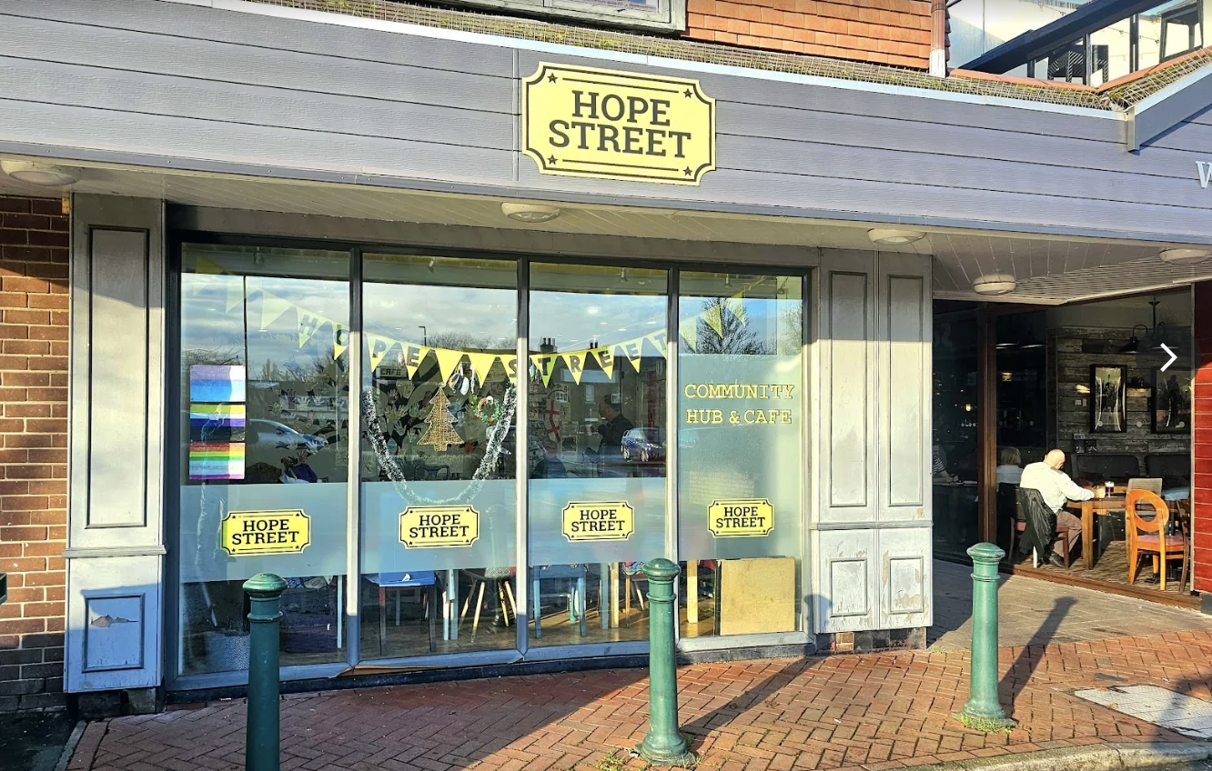 Front of a community hub and cafe named Hope Street with large glass windows decorated with yellow banners, signs, and holiday-themed decorations, and a person sitting inside at a table.