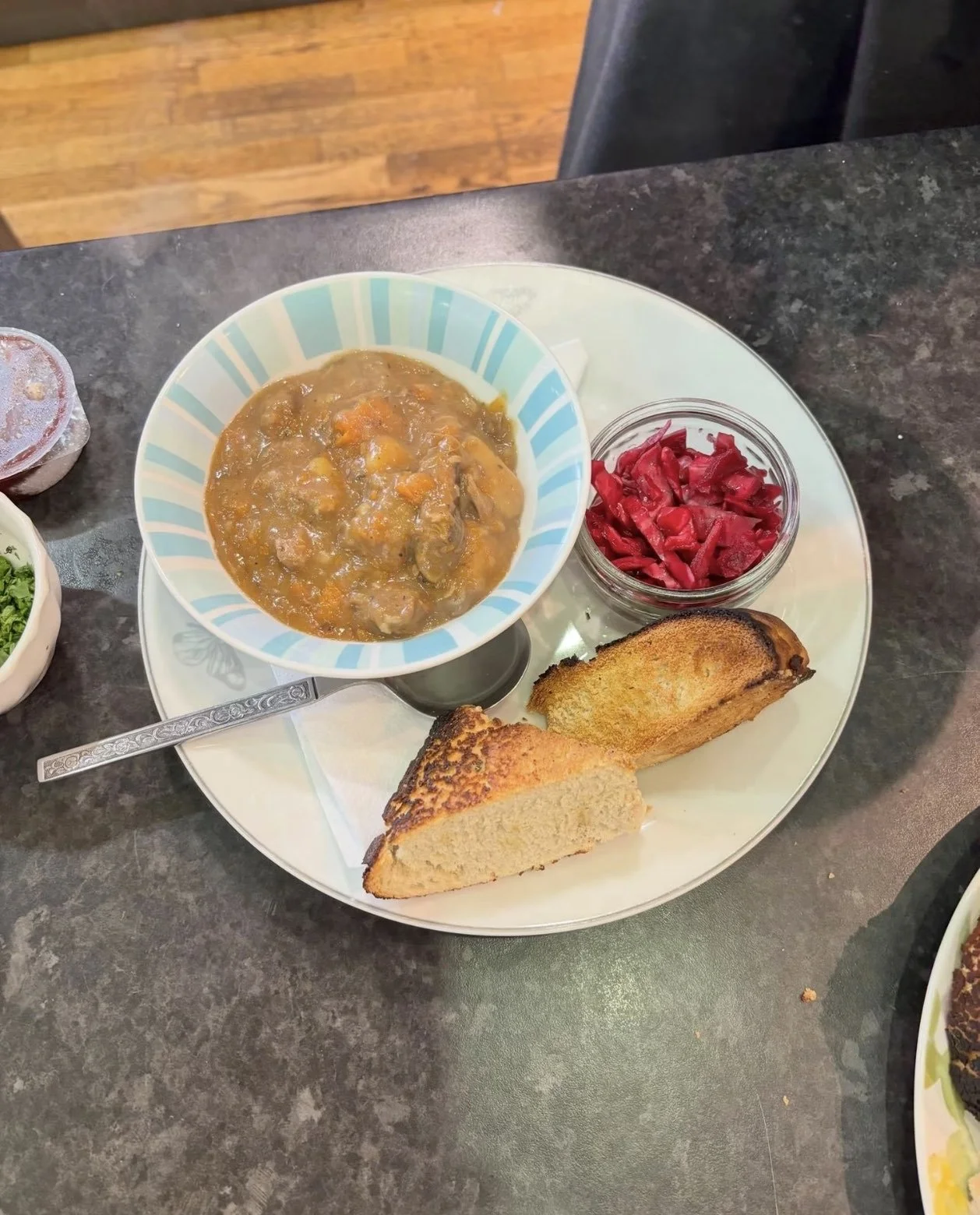 A plate with a bowl of beef and vegetable stew, a small glass jar of pickled beets, and two slices of toasted bread on a white plate.