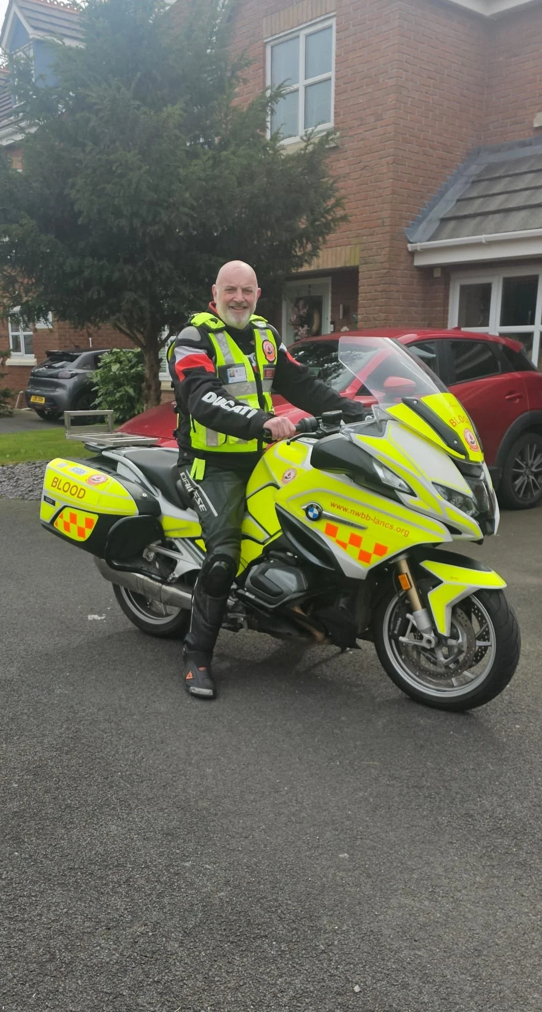 A police officer on a yellow BMW motorcycle is standing on a residential street with houses and parked cars in the background. He is smiling and wearing a black and red police uniform with high-visibility yellow markings.
