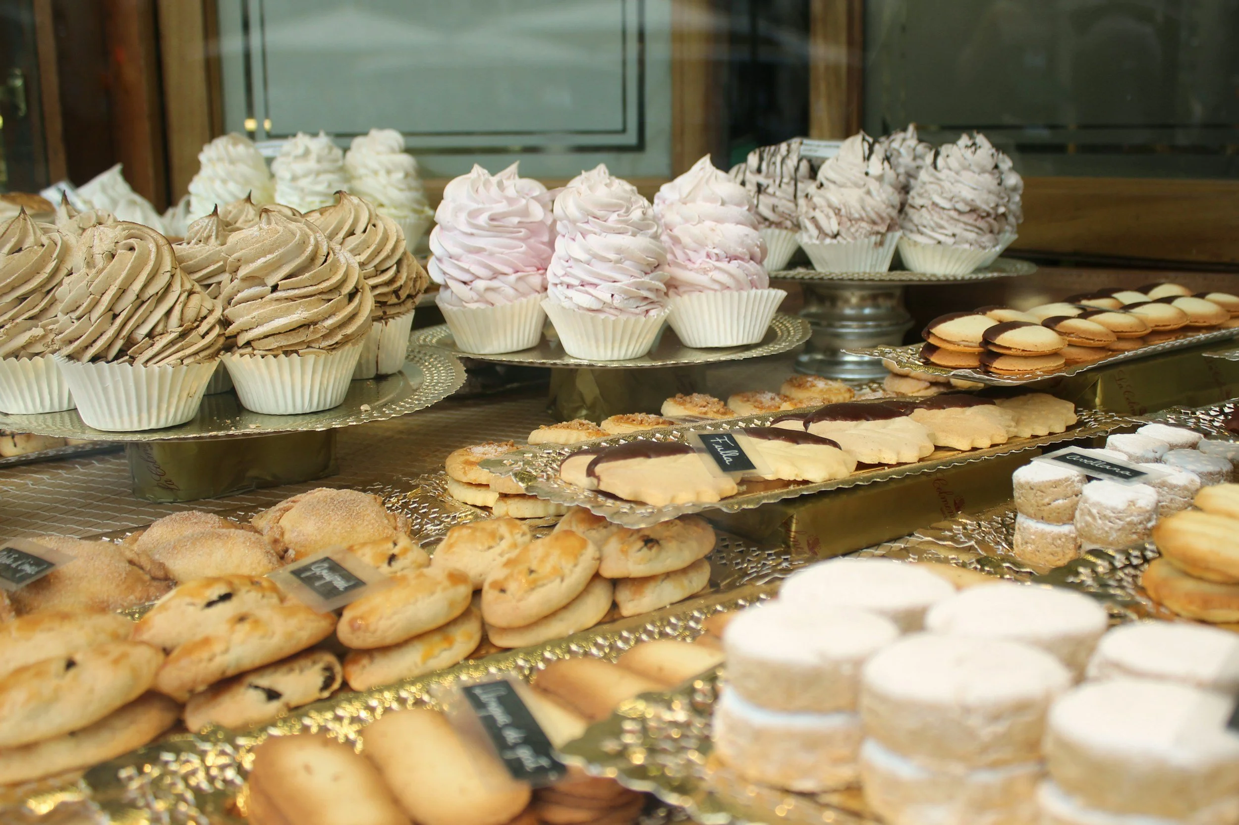 A display of assorted baked goods including muffins, cookies, and macarons on silver trays in a bakery showcase.
