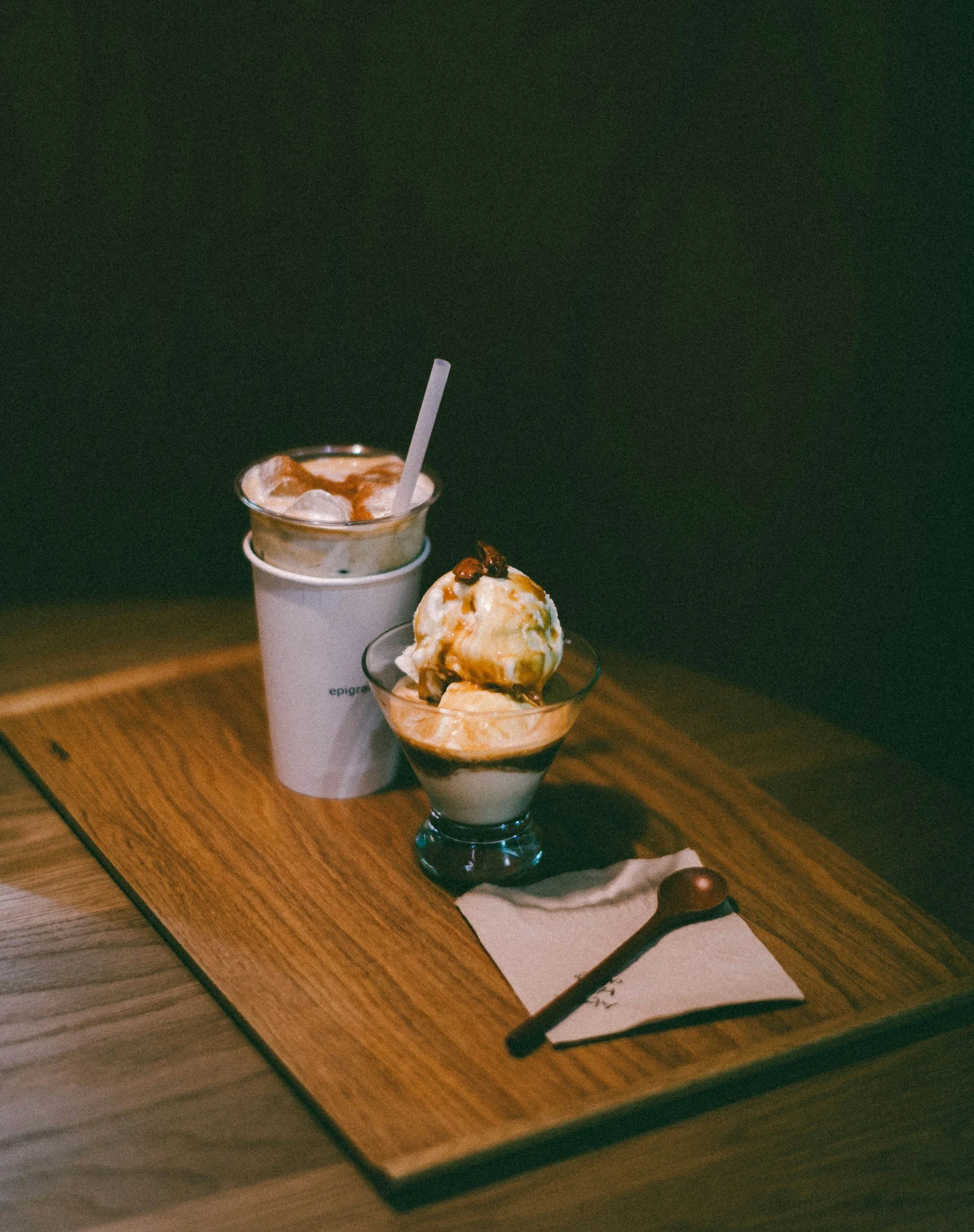 A wooden table with a white napkin, a chocolate spoon, and two dessert drinks: one with ice, a straw, and caramel syrup, and another with ice cream and caramel sauce topped with nuts.