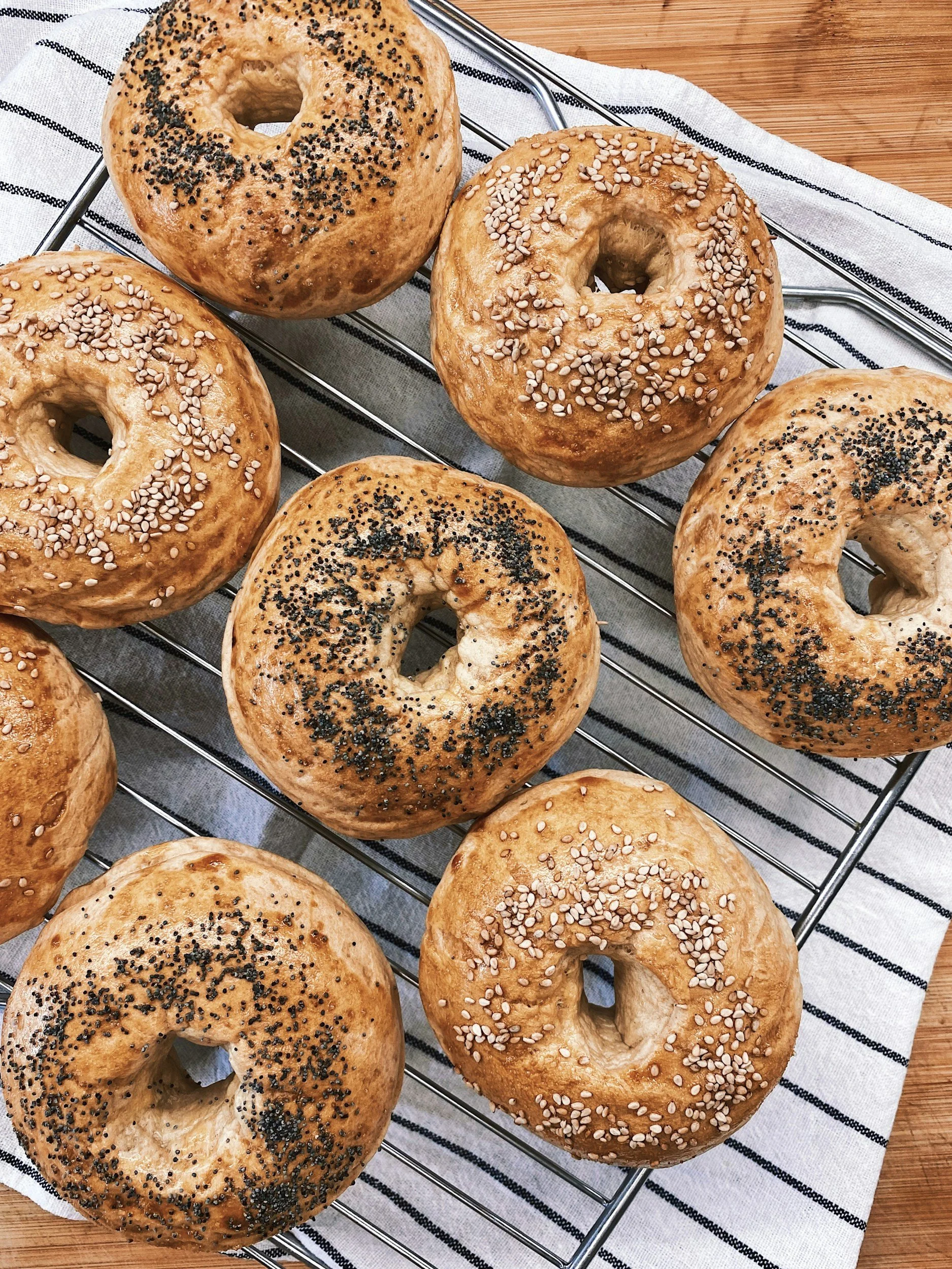 Six bagels on a metal cooling rack, some topped with poppy seeds and others with sesame seeds, placed on a striped cloth on a wooden surface.
