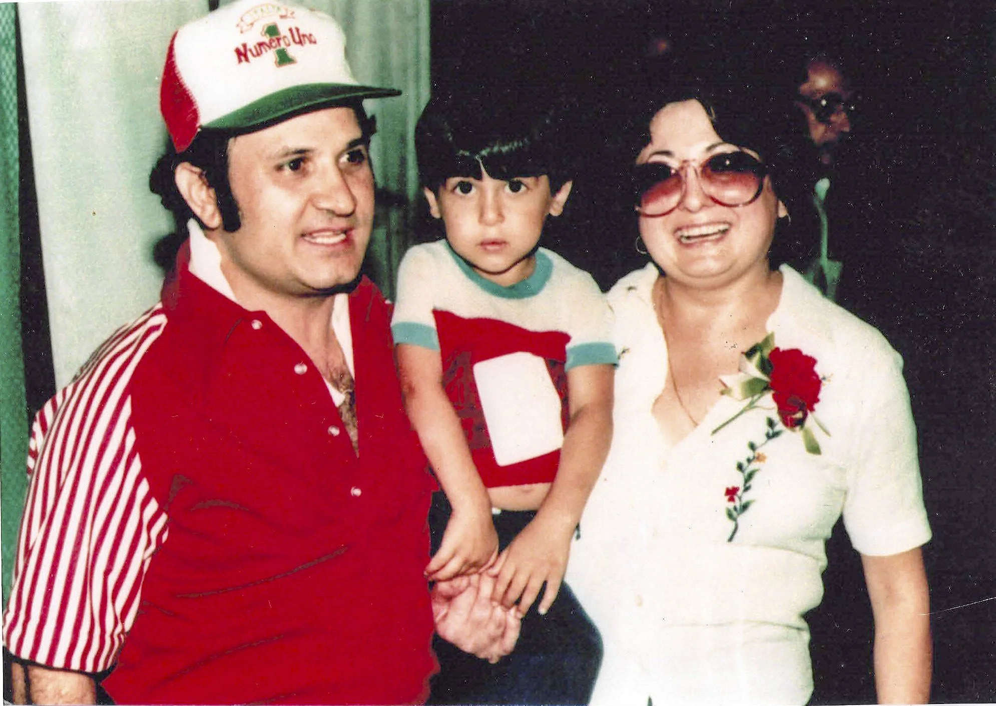 A man, a young boy, and a woman stand close together, holding hands. The man is wearing a red and white striped shirt and a white hat with red and green text. The young boy is wearing a red, white, and teal shirt. The woman is wearing large sunglasses and a white dress with embroidery, holding a red rose.