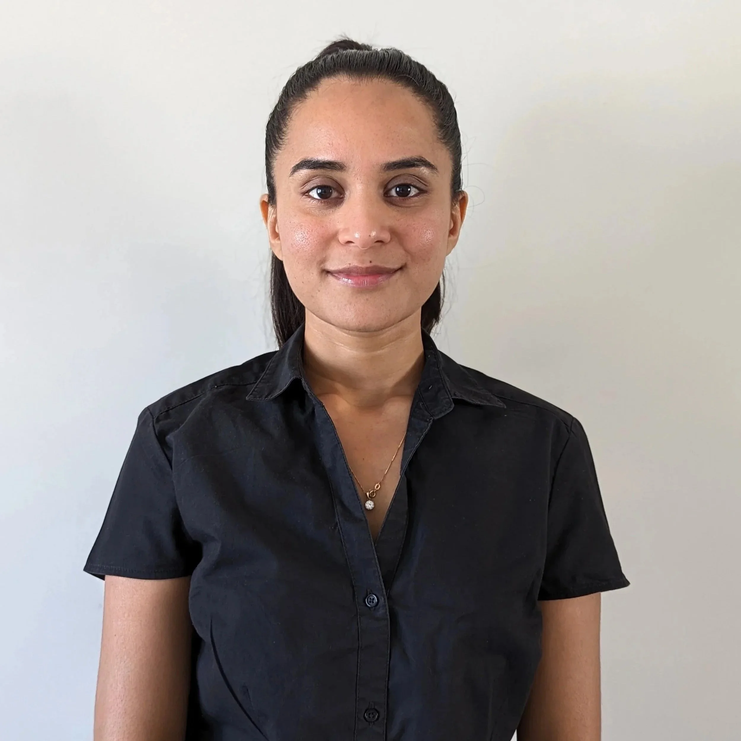 A woman with dark hair tied in a bun, wearing a black button-up shirt and a gold necklace, standing against a plain white wall.