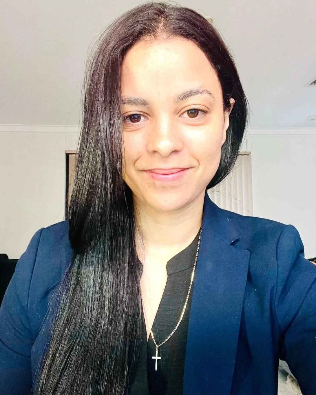 A woman with long black hair and light skin taking a selfie indoors. She is smiling and wearing a dark blazer, a black top, and a silver cross necklace.