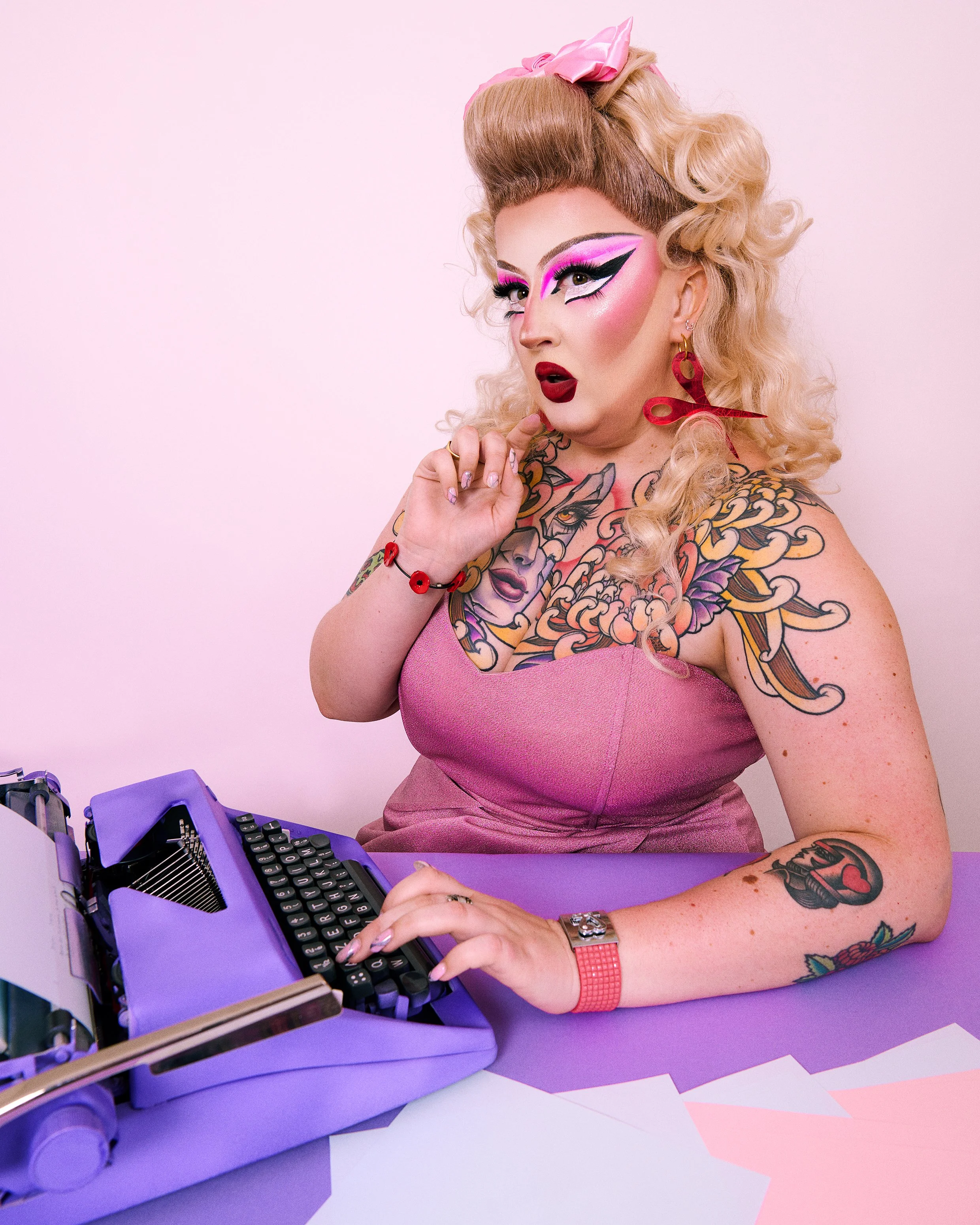Angelica est habillée d'une robe rose avec un maquillage drag et des tatouages apparants, utilisant une machine à écrire violette.