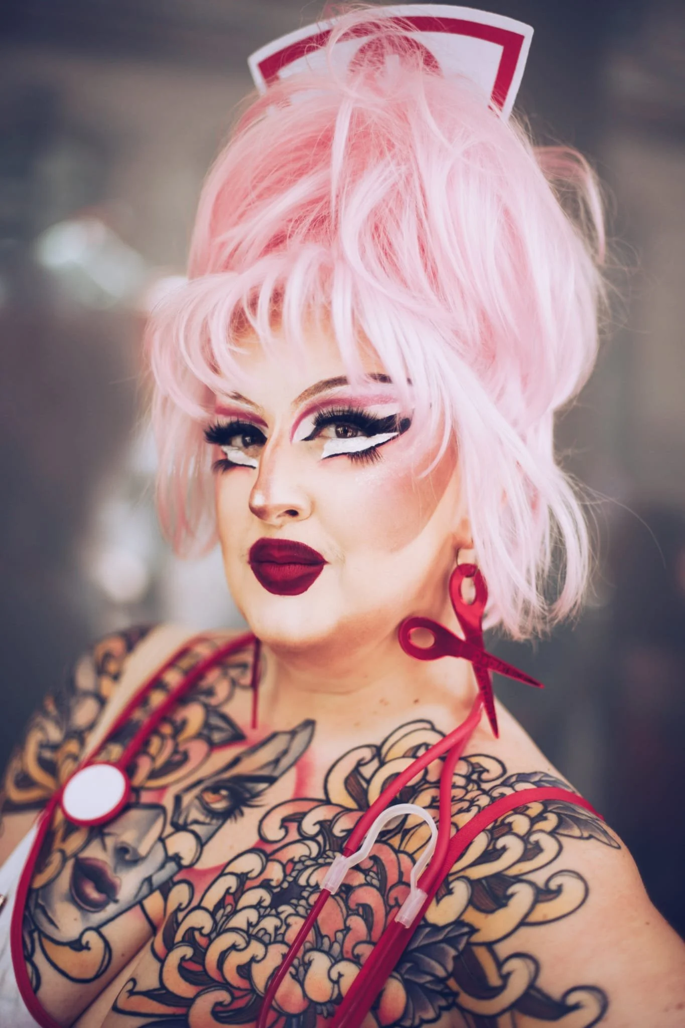 Angelica Stratrice avec des cheveux roses, maquillage drag, tatouages sur le bras et un stéthoscope rouge autour du cou.