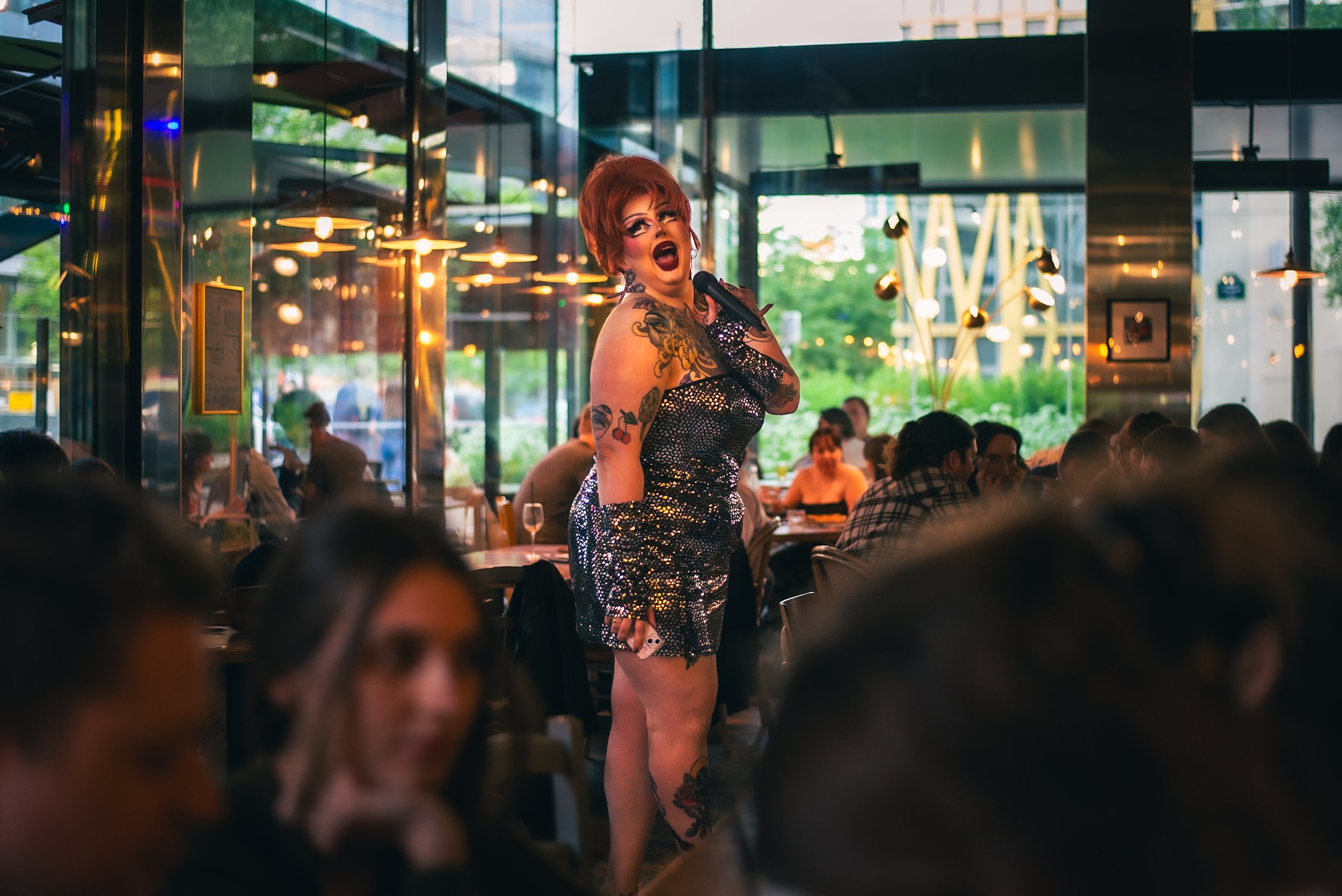 Angelica Stratrice porte un micro, animant la soirée du restaurant La Birreria, entourée de personnes assises à des tables, dans un décor moderne et lumineux avec des lumières chaudes