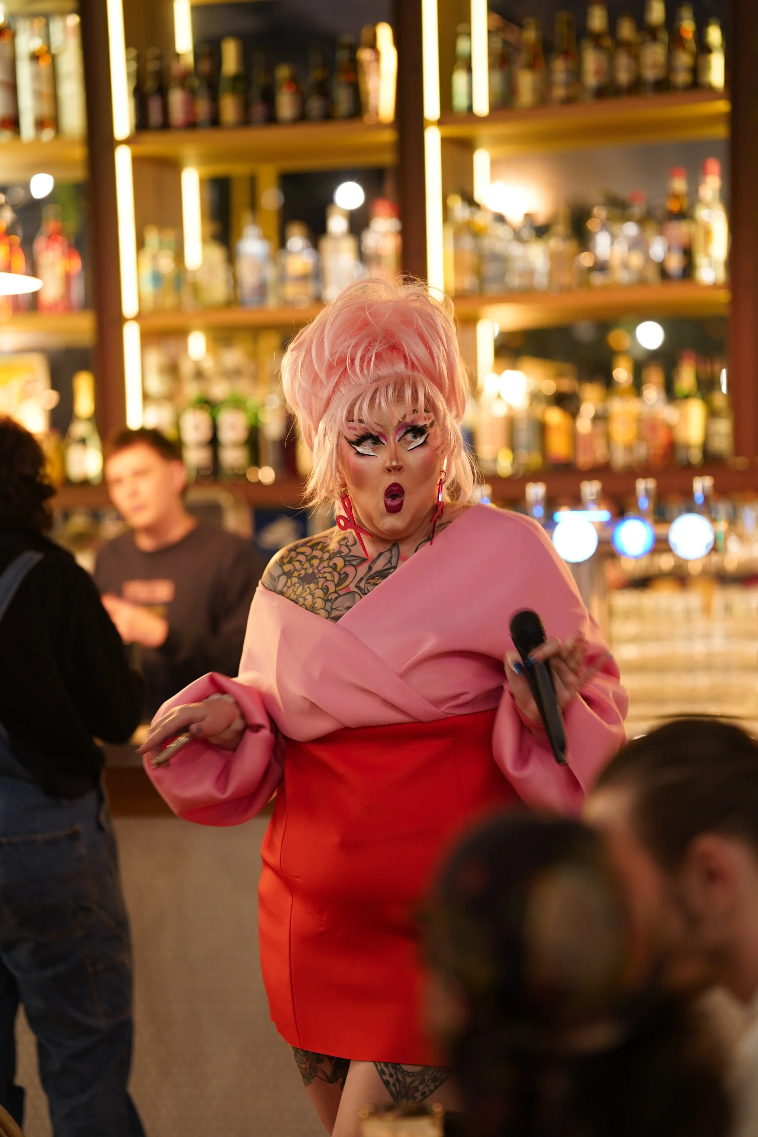 Angelica Stratrice, ici avec des cheveux roses, maquillage élaboré, portant une robe rose et rouge, tenant un micro, dans un bar avec des étagères de bouteilles en arrière-plan.