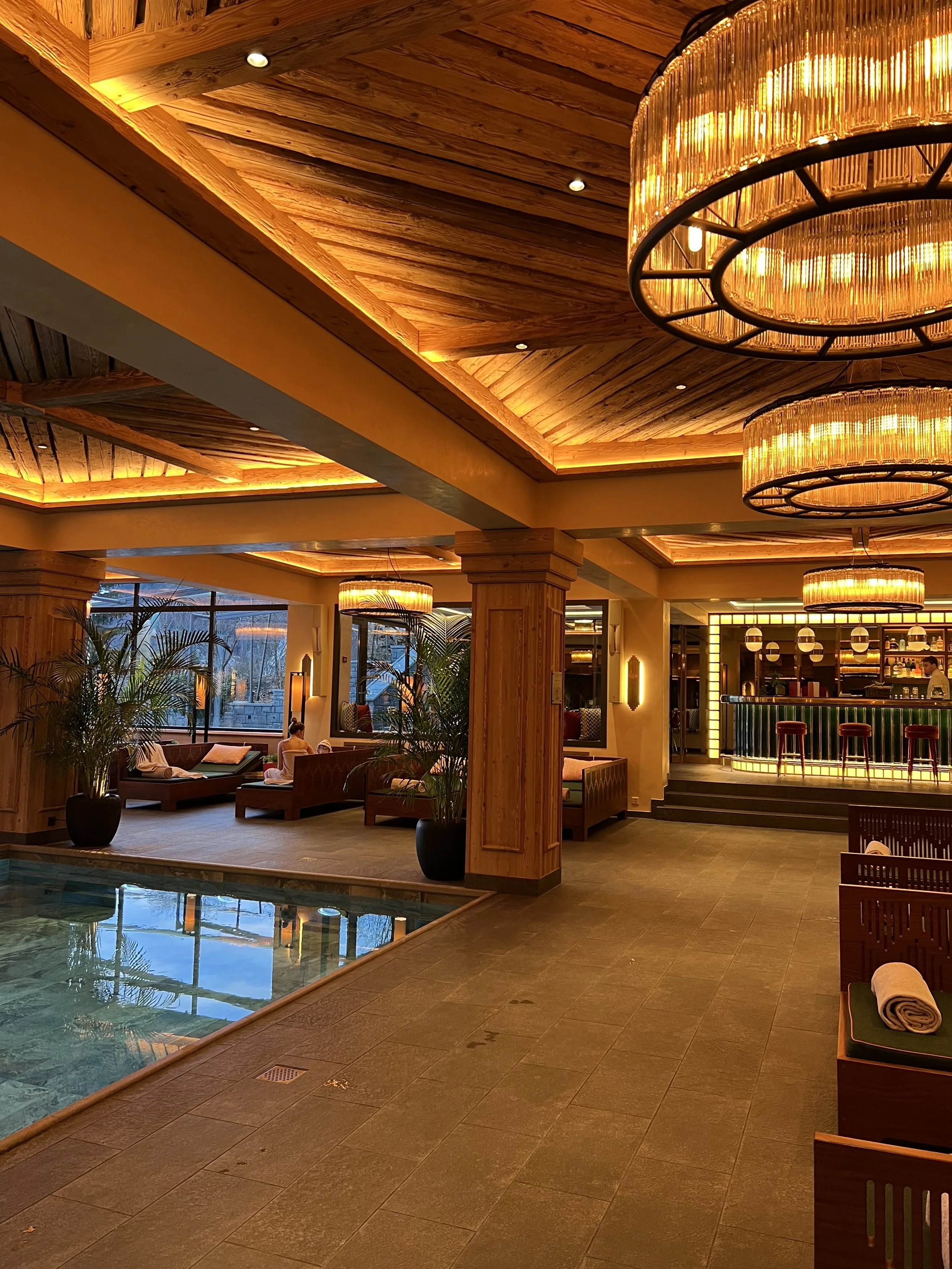 An upscale hotel lobby with wooden ceiling and warm lighting, including large chandeliers, seating area with lounge chairs, potted plants, and a swimming pool visible through a window.