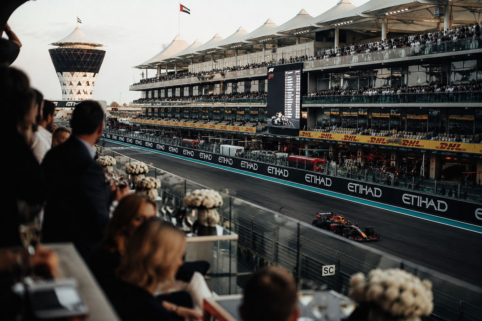 View of a Formula 1 race at the Abu Dhabi Grand Prix with cars on the track, spectators in the grandstands, and team members on the pit lane. The scene is set at sunset with a large tower in the background.