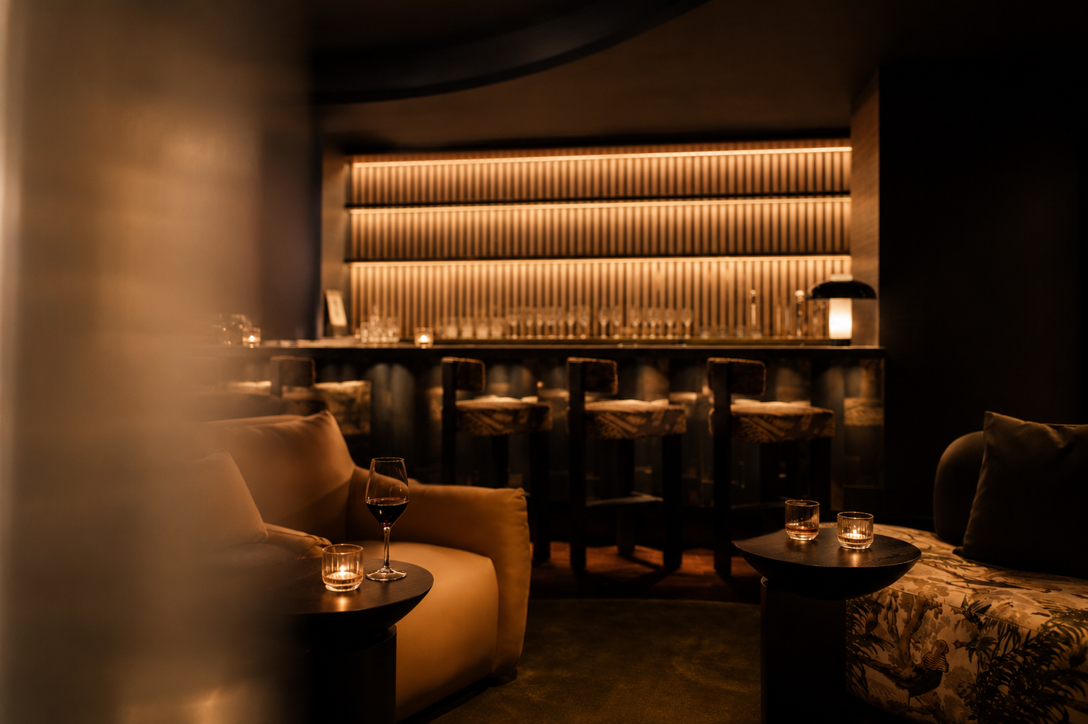 A cozy bar area with dim lighting, featuring a bar counter with barstools, a sofa with a glass of red wine and candles, and soft lighting on the architectural wall behind the bar.