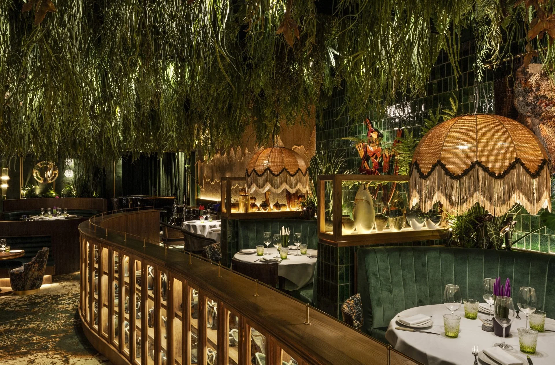 Elegant restaurant interior with lush green hanging plants and dim lighting, featuring tables set for dining with white tablecloths, glassware, and decorative lamps with fringed shades.