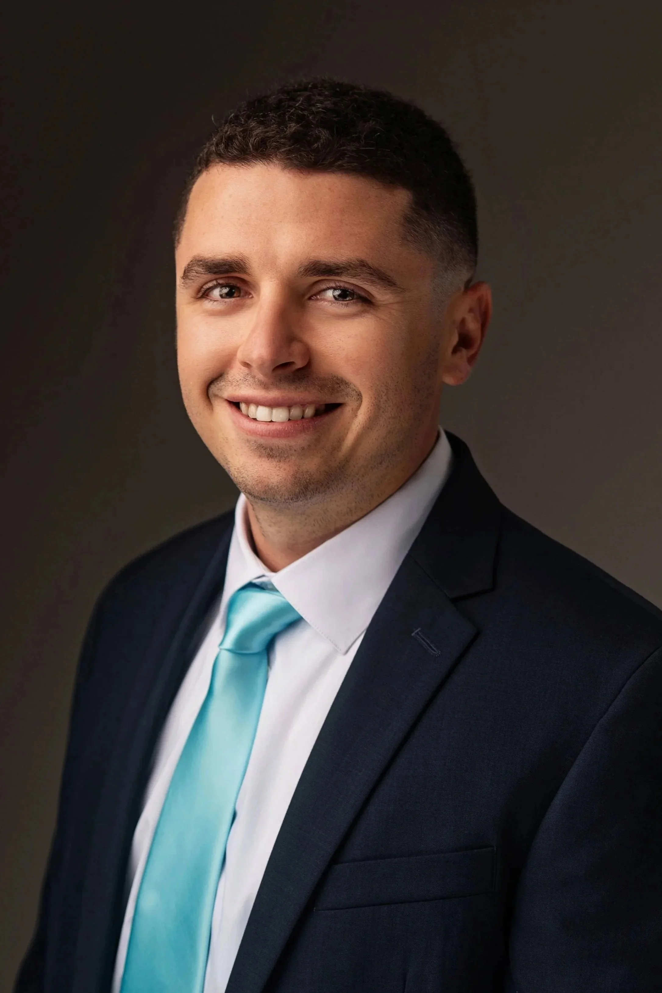 Professional portrait of a smiling man with short dark hair, wearing a dark suit, white shirt, and turquoise tie against a dark background.