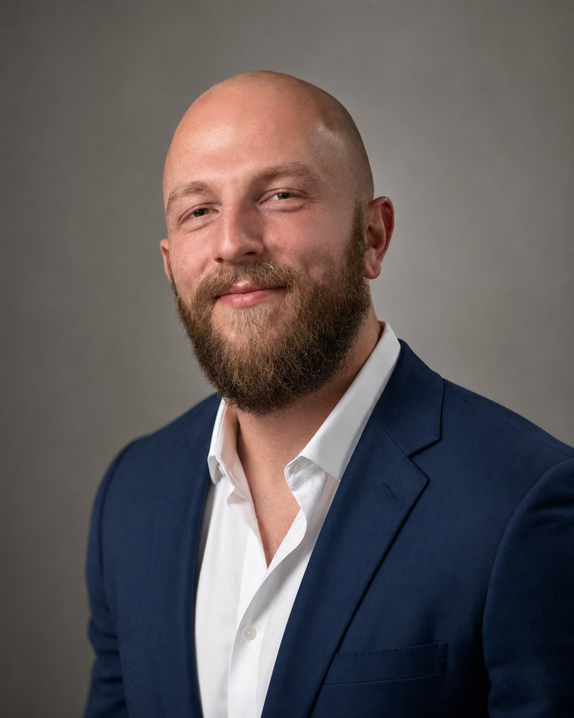 Headshot of a man with a beard and bald head, wearing a navy blazer and white shirt, smiling against a gray background.