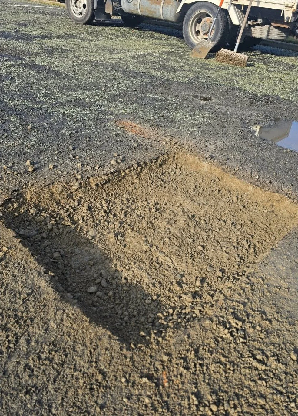 A dug-out rectangular hole in a gravel surface near a parked utility truck, with a broom and a shovel leaning against the truck.