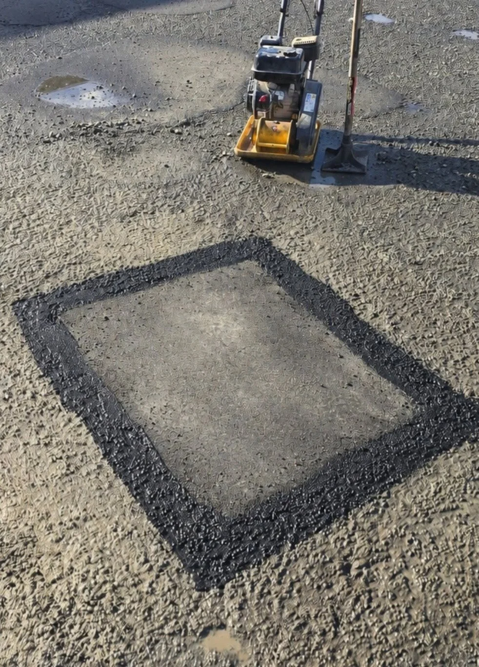 Square outline in freshly laid asphalt, construction tools nearby, on an asphalt surface.