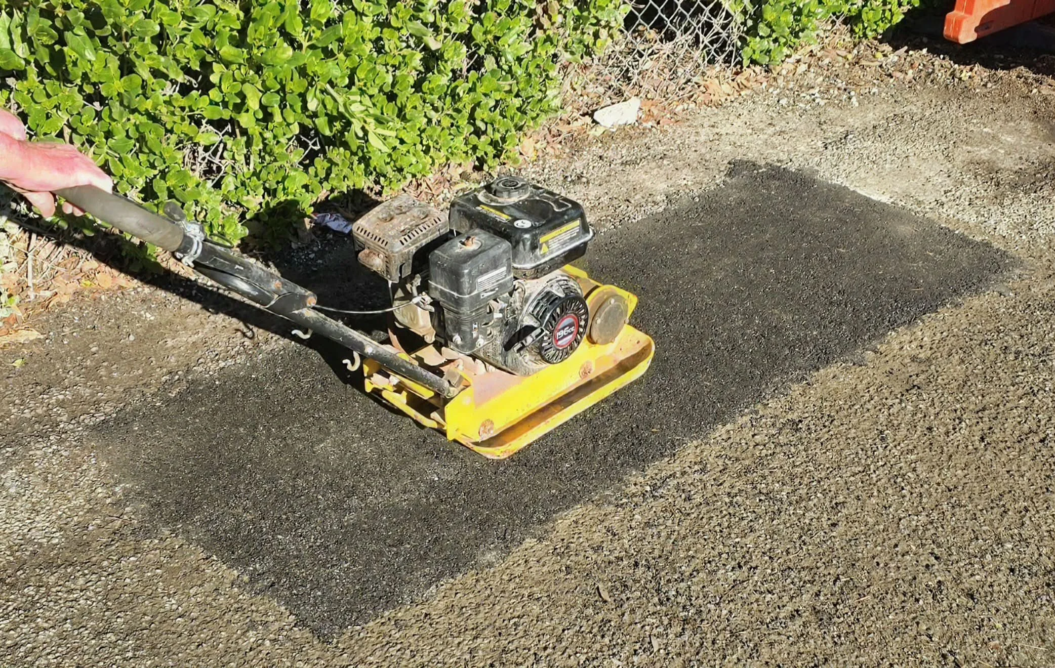 A person using a plate compactor to level a gravel or dirt surface outdoors, with a green hedge and a chain-link fence in the background.
