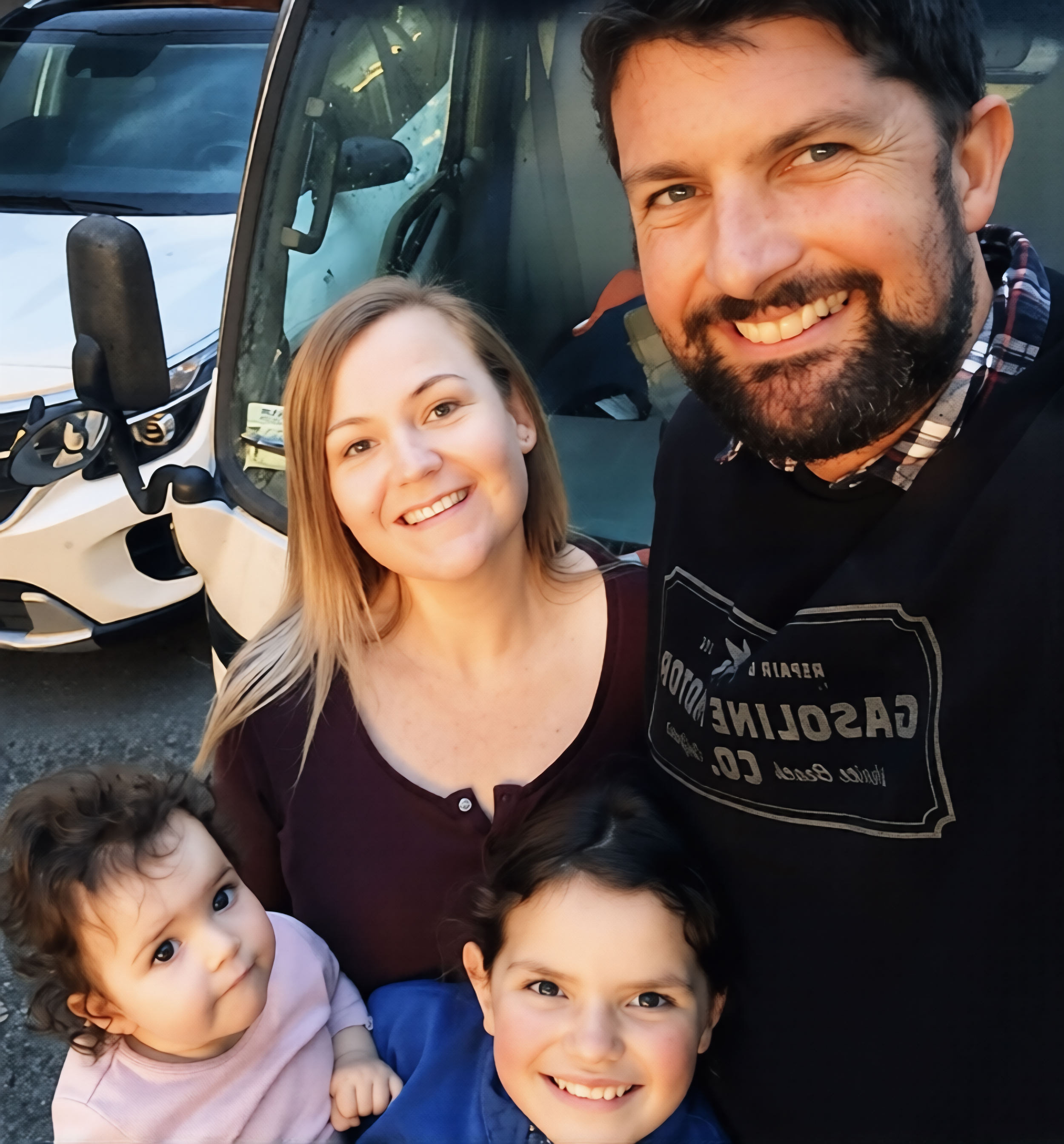 A smiling man with a beard, a woman with blonde hair, and two children with dark hair and eyes, outside next to a white vehicle.