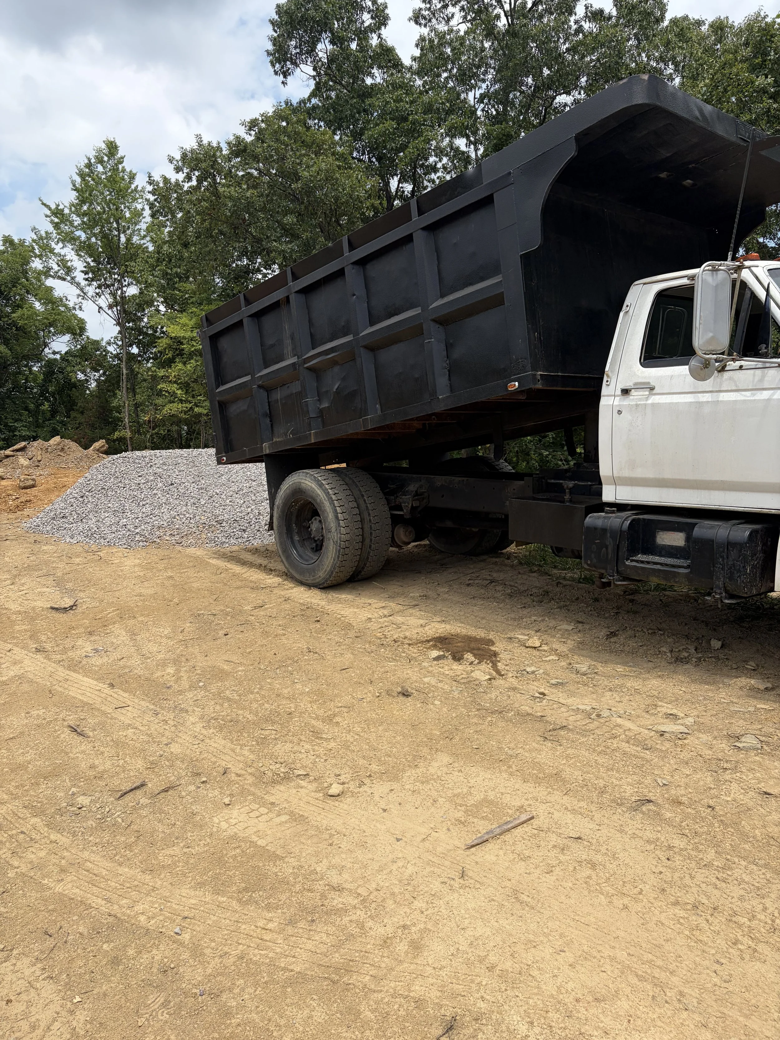 A large white dump truck delivering rock near Morristown Tennessee with a black container is parked on a dirt area, with a pile of gravel behind it and green trees in the background.