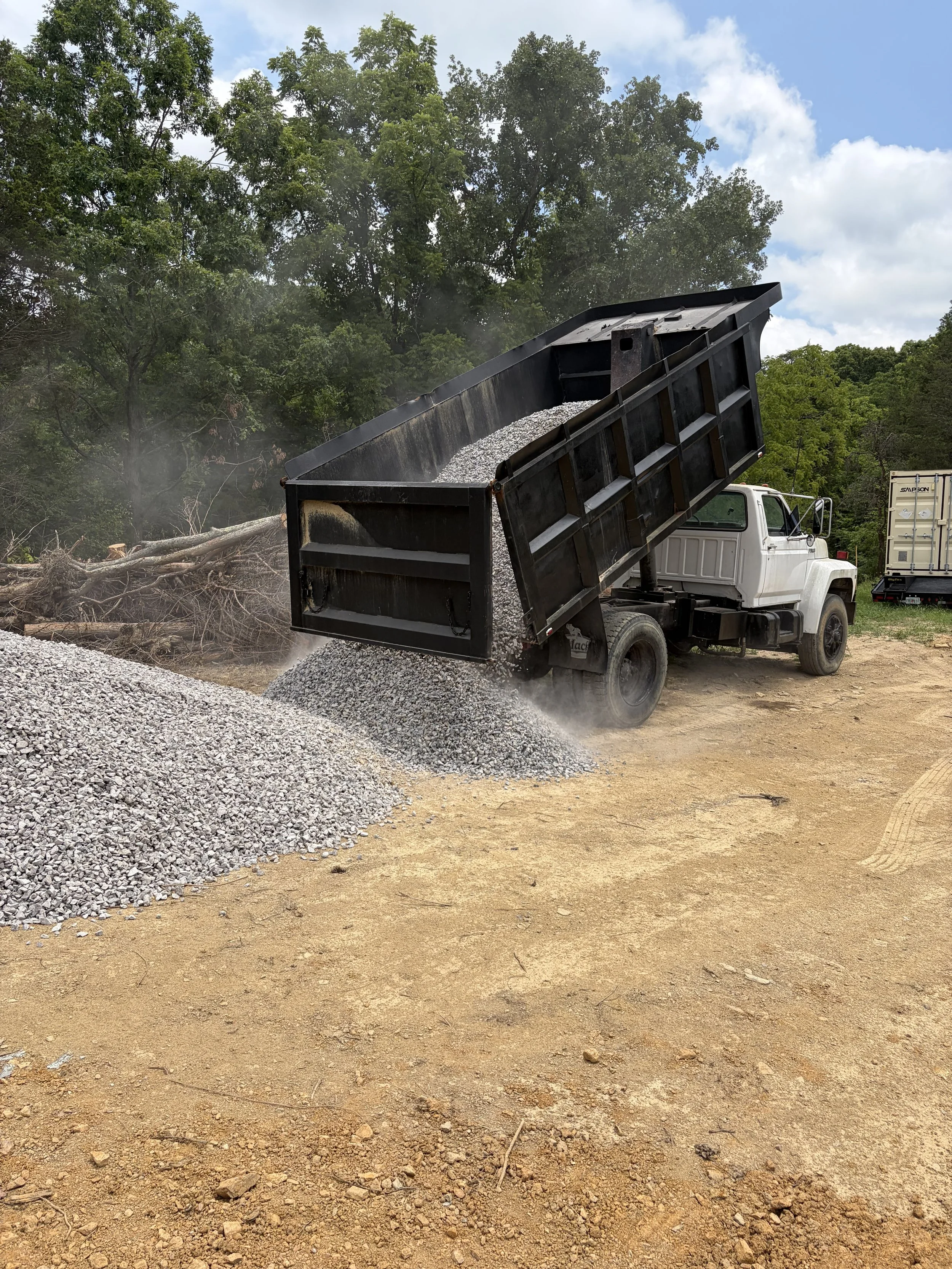 A dump truck with a black bed is unloading gravel onto a dirt surface at a construction site, with trees and a partly cloudy sky in the background in Rogersville Tennessee.