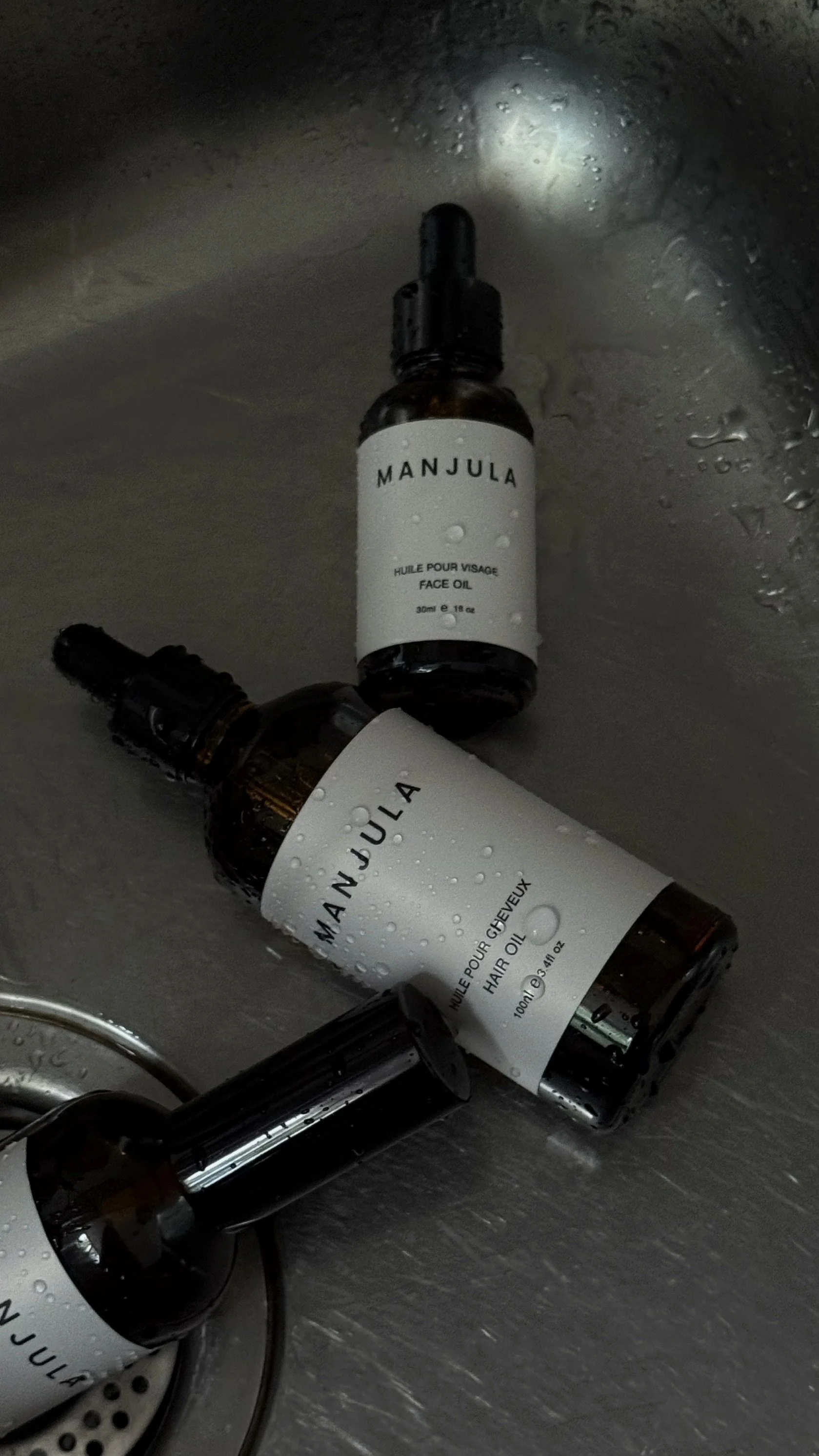 Three bottles of Manjula face and hair oil are lying on a metallic surface with water droplets on them.