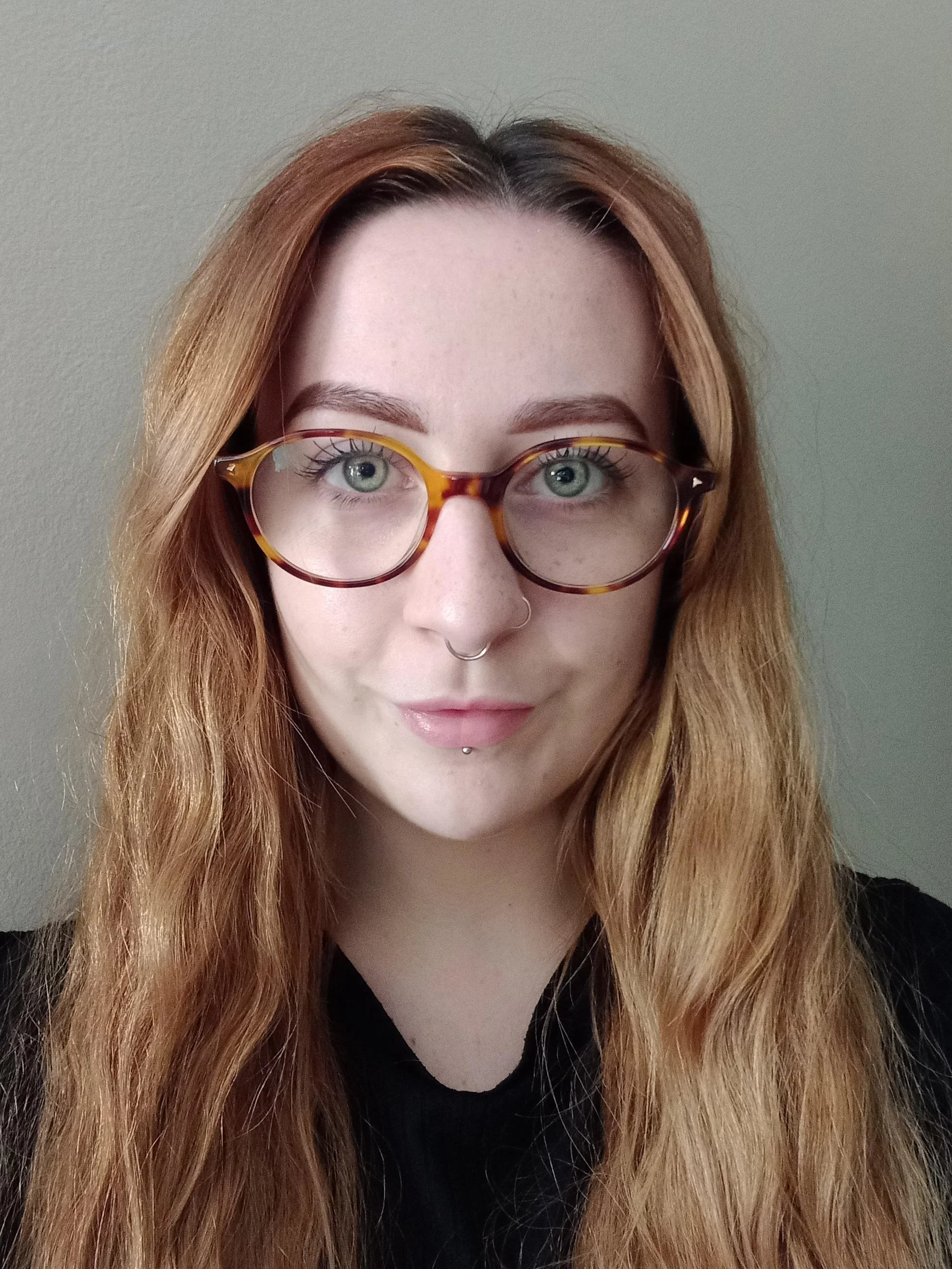 A woman with long, wavy red hair wearing large, tortoiseshell glasses and a septum piercing, looking directly at the camera, against a plain, light-colored background.