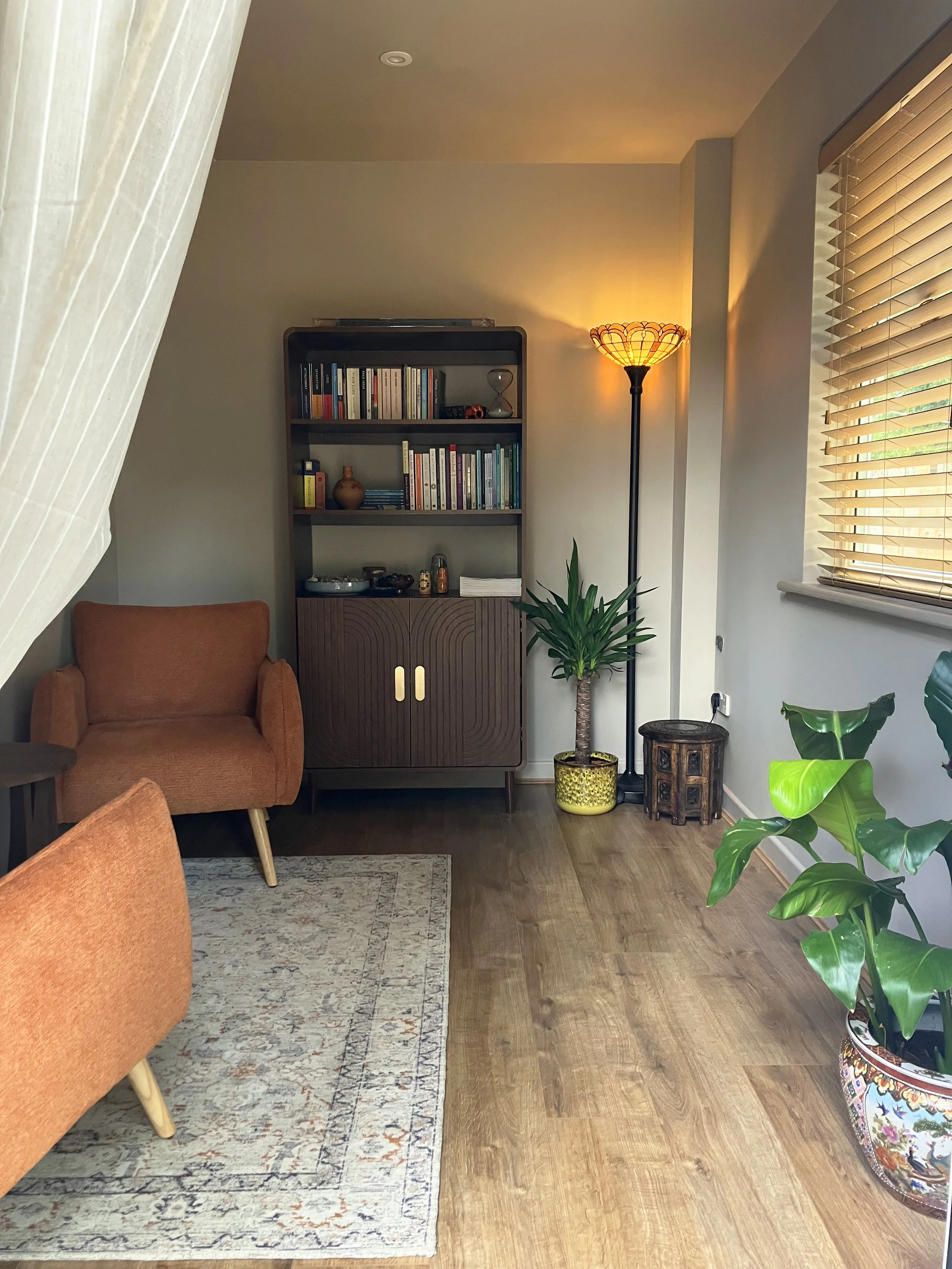 A cozy living room corner with a brown upholstered armchair on the left, a patterned area rug, a wooden bookshelf filled with books and decorative items, a tall floor lamp with a warm-colored shade, a window with wooden blinds, and a large potted plant with broad green leaves.