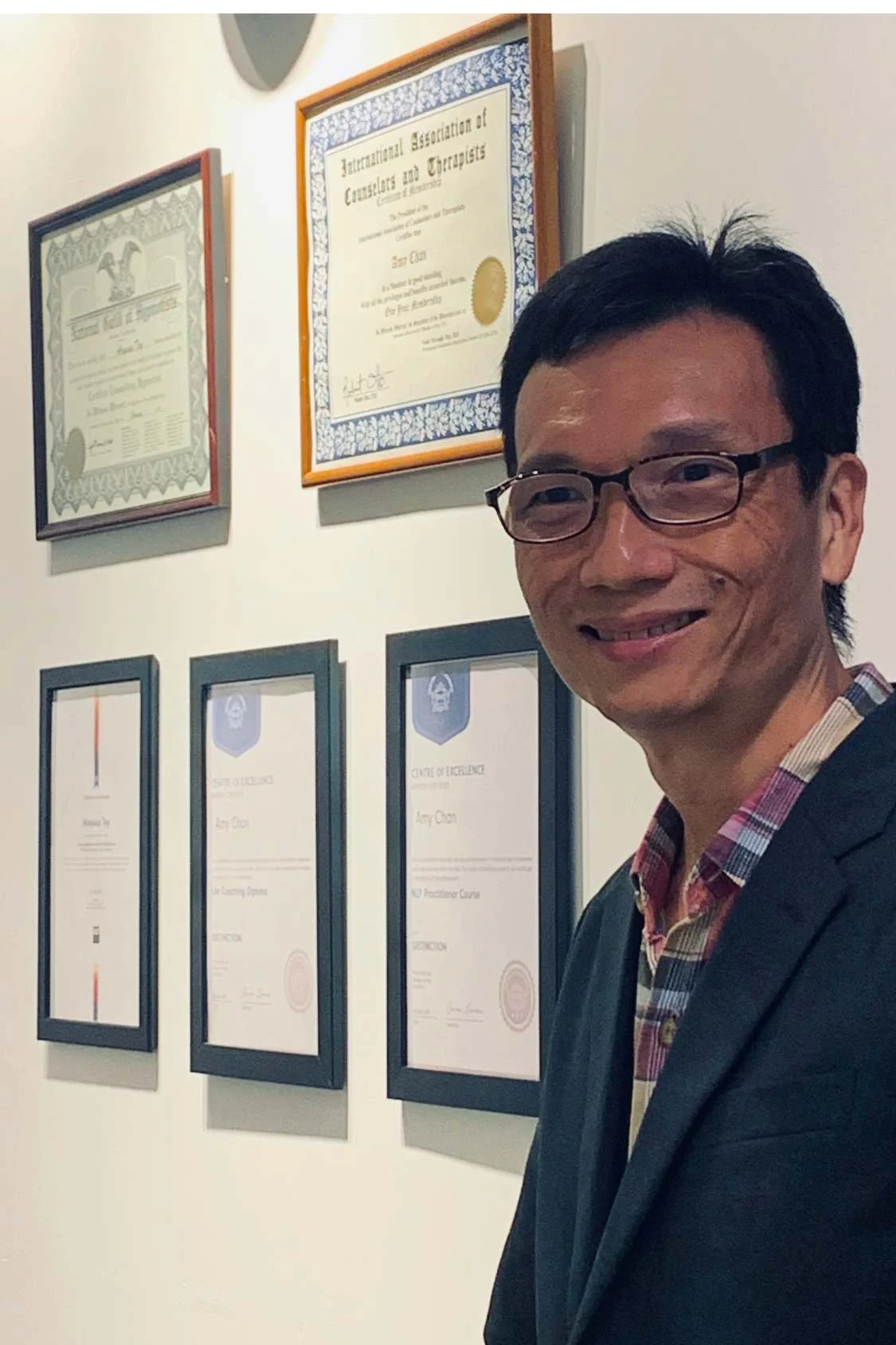 A man wearing glasses and a blazer standing in front of a wall with framed certificates and awards.