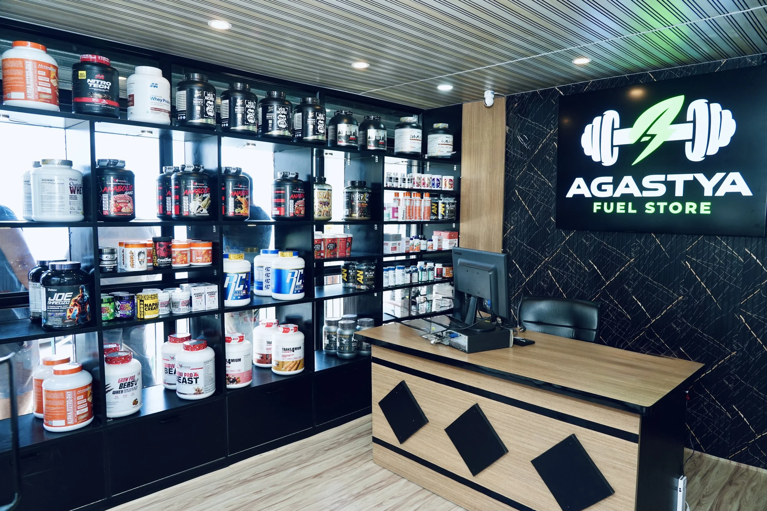 Inside a fuel store with shelves stocked with various protein supplements, energy drinks, and fitness products. A neon sign reads 'AGASTYA Fuel Store' and features a logo with a dumbbell, lightning bolt, and green leaf.