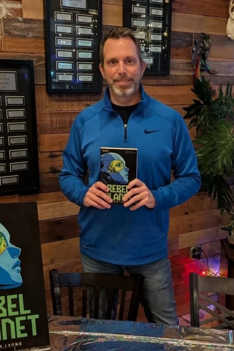 A man with a beard and mustache wearing a blue Nike jacket is holding a book titled 'Rebel Mother' in a room with wooden walls and framed awards or certificates behind him. There is a plant to his right and a painting of a woman in blue with goggles and a yellow eye patch on the table in front of him.