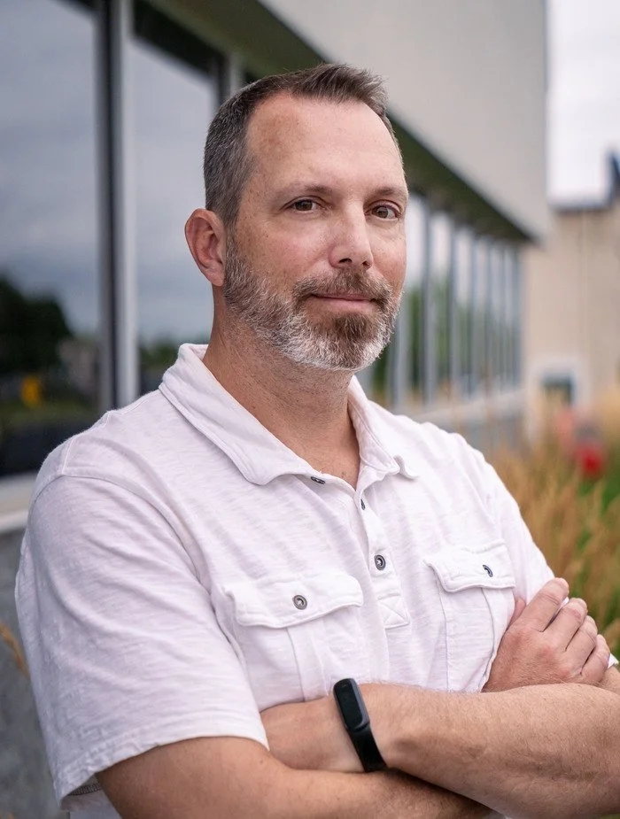 A middle-aged man with short dark hair, a beard, and a mustache stands outdoors with arms crossed, wearing a white short-sleeved button-up shirt and a black smartwatch, in front of a building with large windows and some plants.