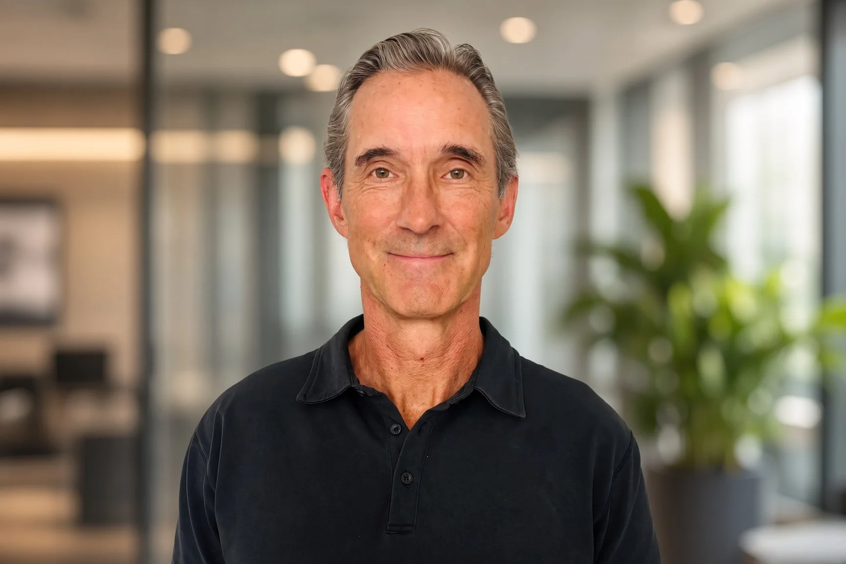 A middle-aged man with gray hair and a friendly expression, wearing a black collared shirt, standing in a modern, well-lit office with blurred background including large windows and green plants.