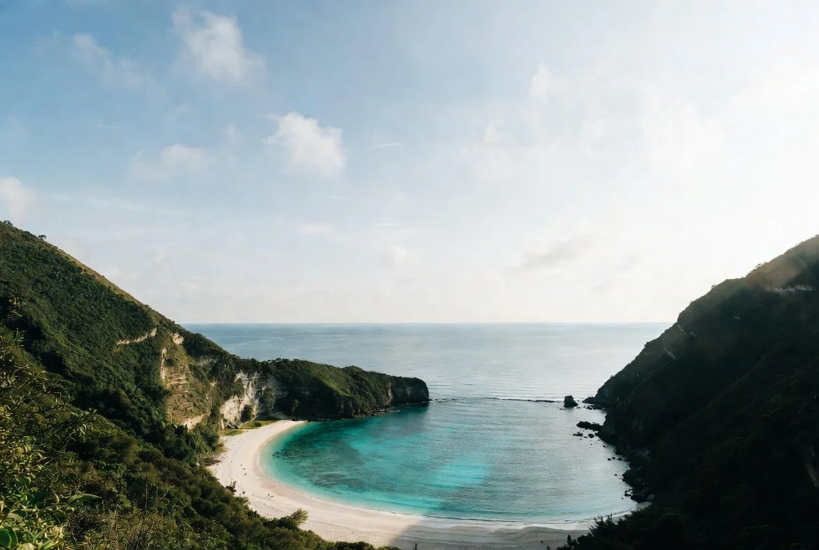A scenic view of a crescent-shaped beach with clear turquoise water, surrounded by green hills on both sides, under a partly cloudy sky.