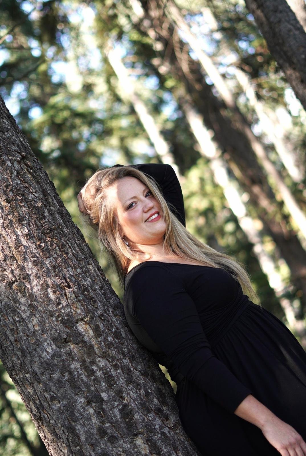 Une femme souriante en robe noire, appuyée contre un arbre dans un environnement forestier. Elle a de longs cheveux blonds et porte un maquillage léger.